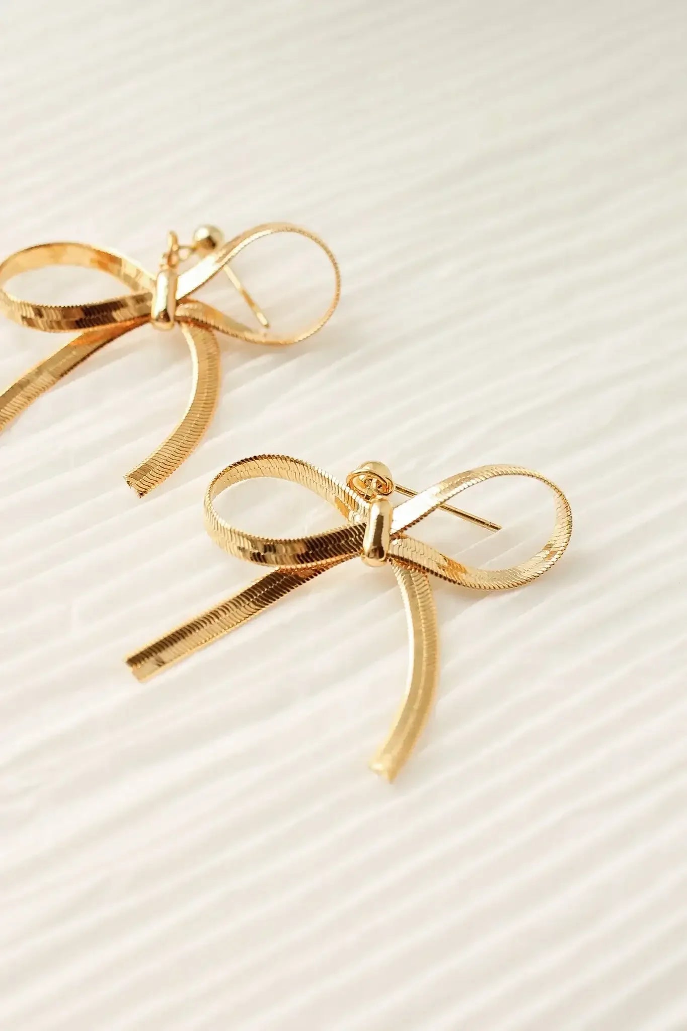 Gold bow-shaped earrings rest on a textured, light-colored surface, showcasing their intricate design and sheen. They lie side by side, emphasizing their decorative style.