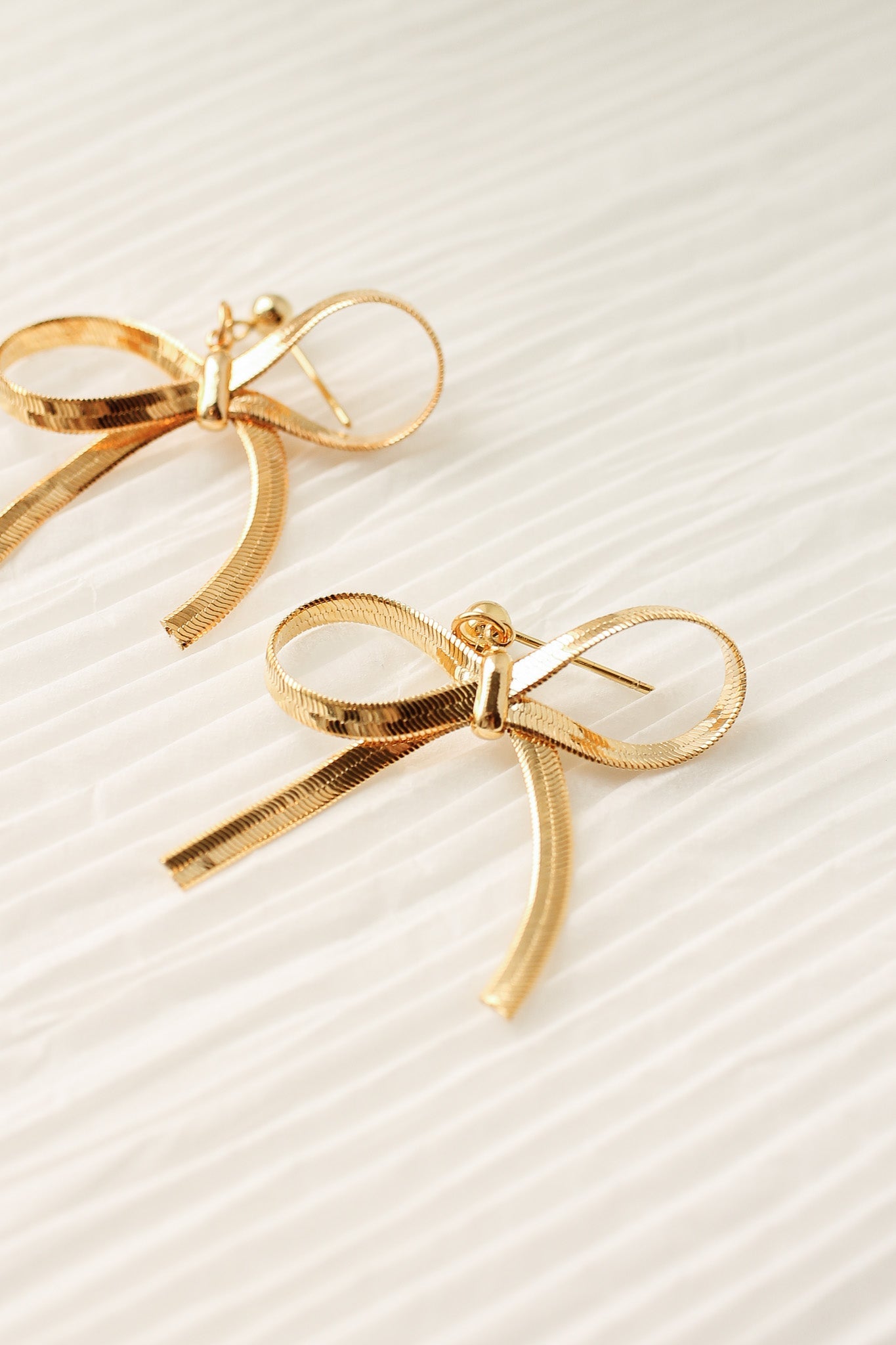 Gold bow-shaped earrings rest on a textured, light-colored surface, showcasing their intricate design and sheen. They lie side by side, emphasizing their decorative style.
