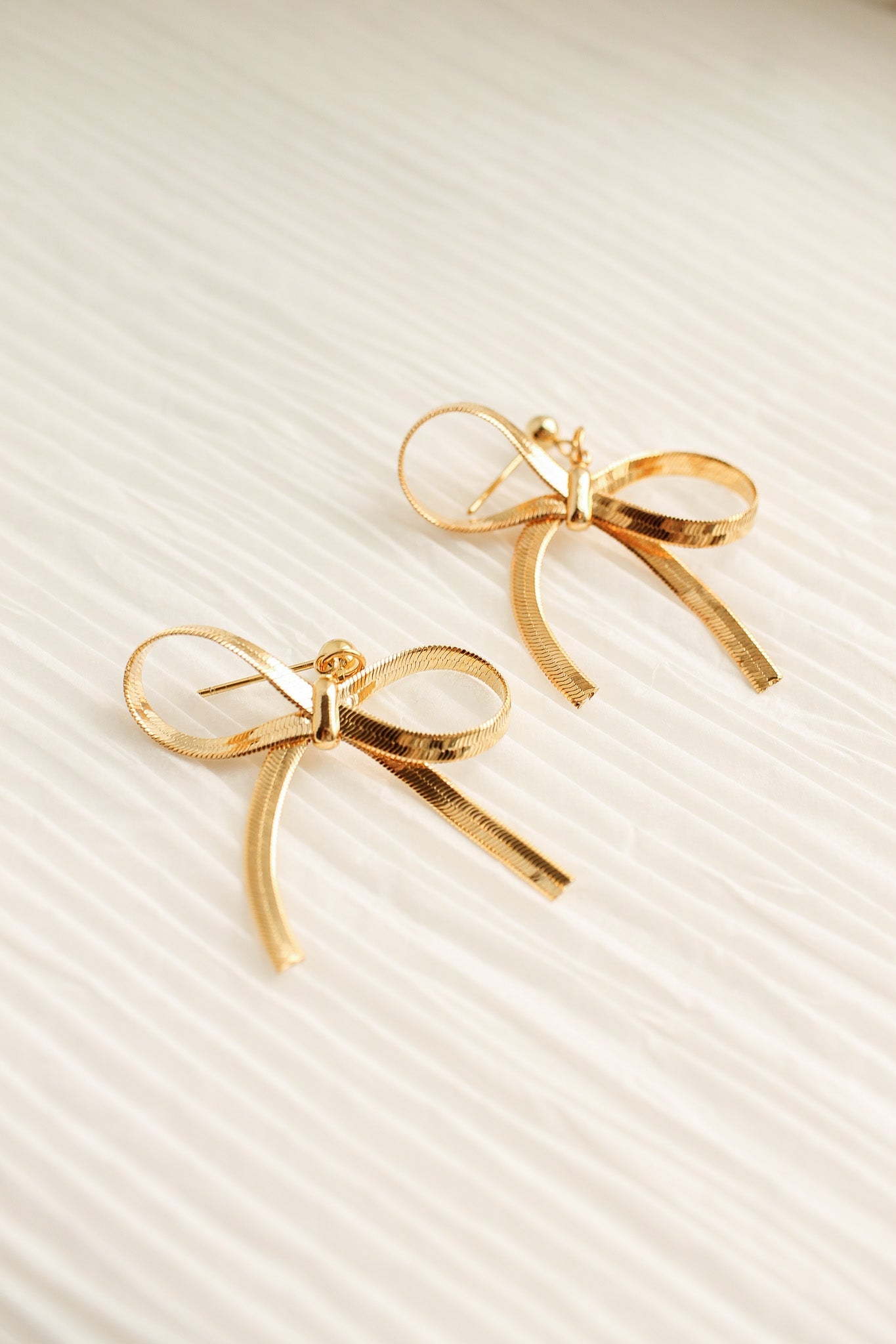 Two golden bow-shaped earrings rest on a soft, textured surface. Their design features loops and tails, reflecting light elegantly, suggesting a delicate and stylish accessory.