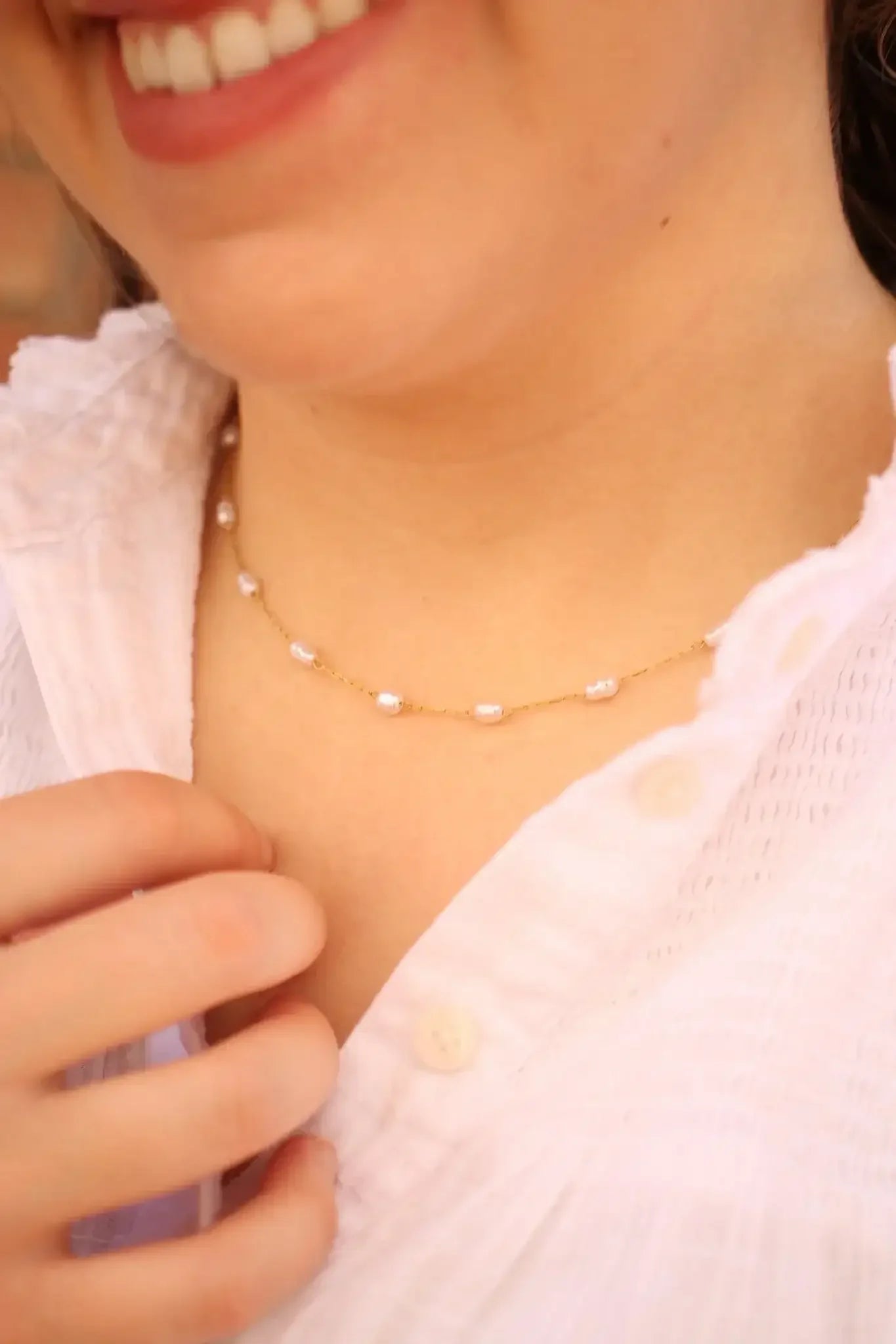 A necklace made of small pearls on a delicate gold chain is worn around a person's neck. The individual gently touches the necklace while wearing a light, button-up blouse.