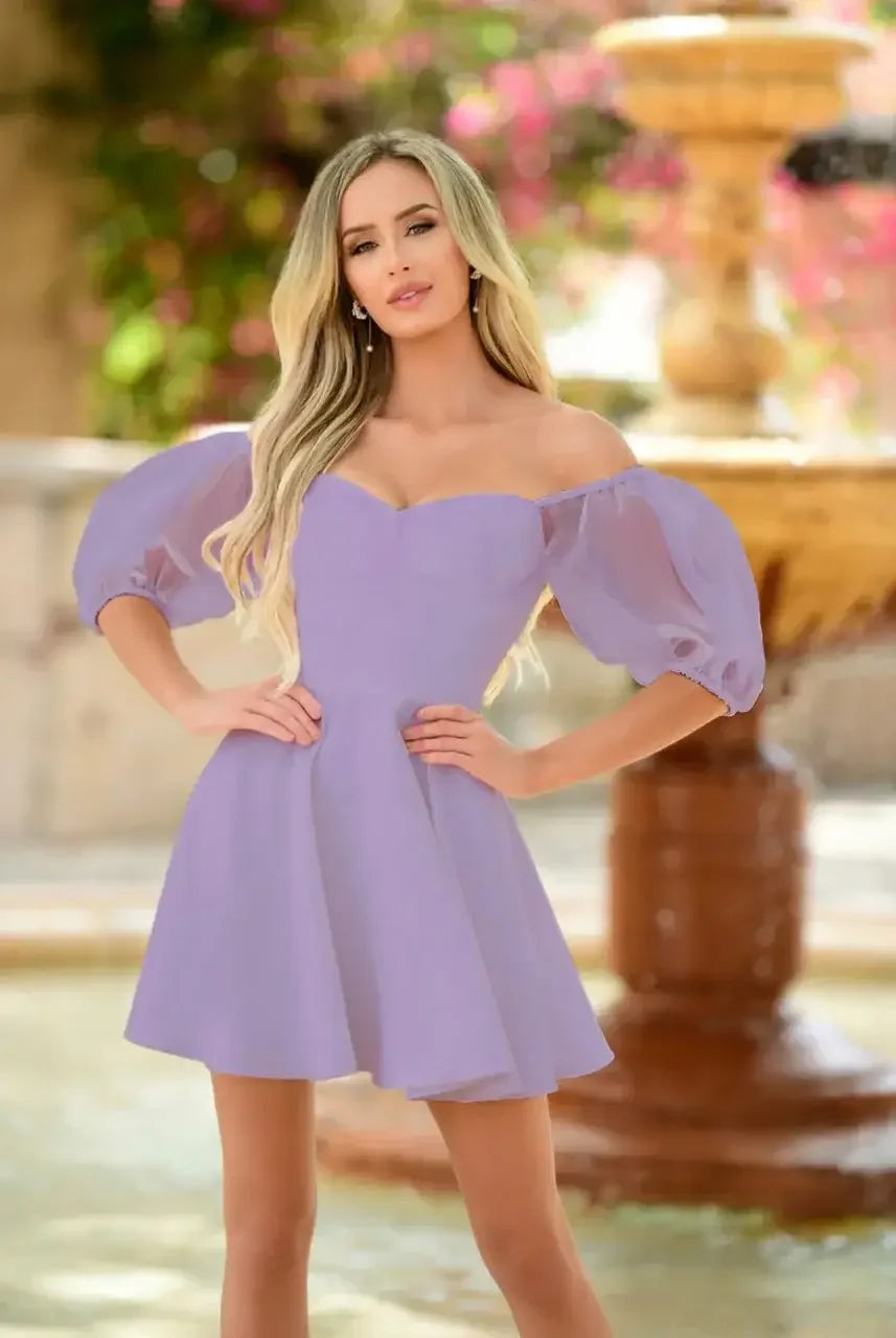 A woman in a lilac off-shoulder dress poses confidently with her hands on her hips, set against a blurred background featuring a fountain and vibrant foliage.