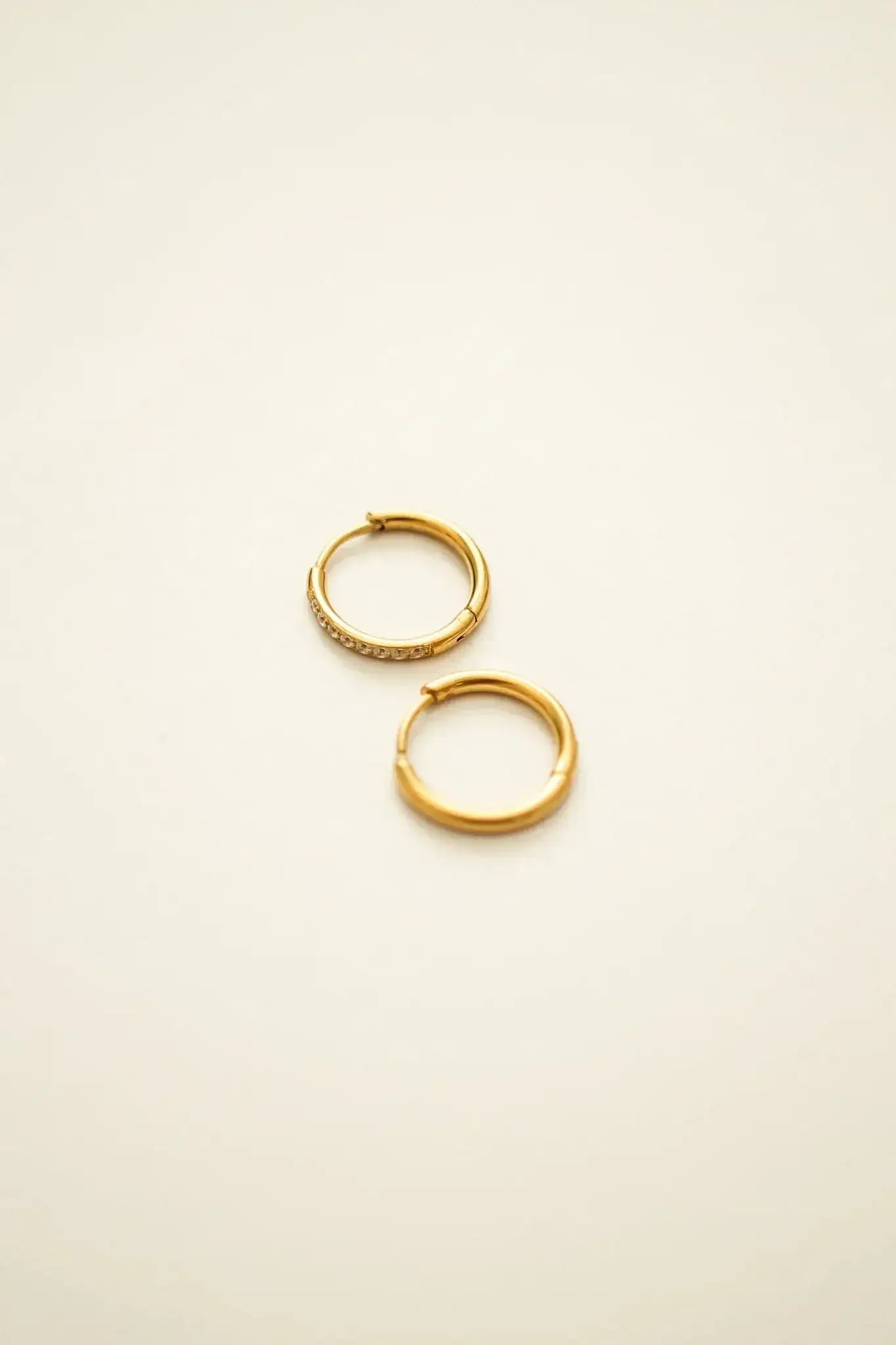 Two gold hoop earrings, one adorned with small sparkling stones, rest on a smooth, light-colored background, highlighting their elegant design and beauty.