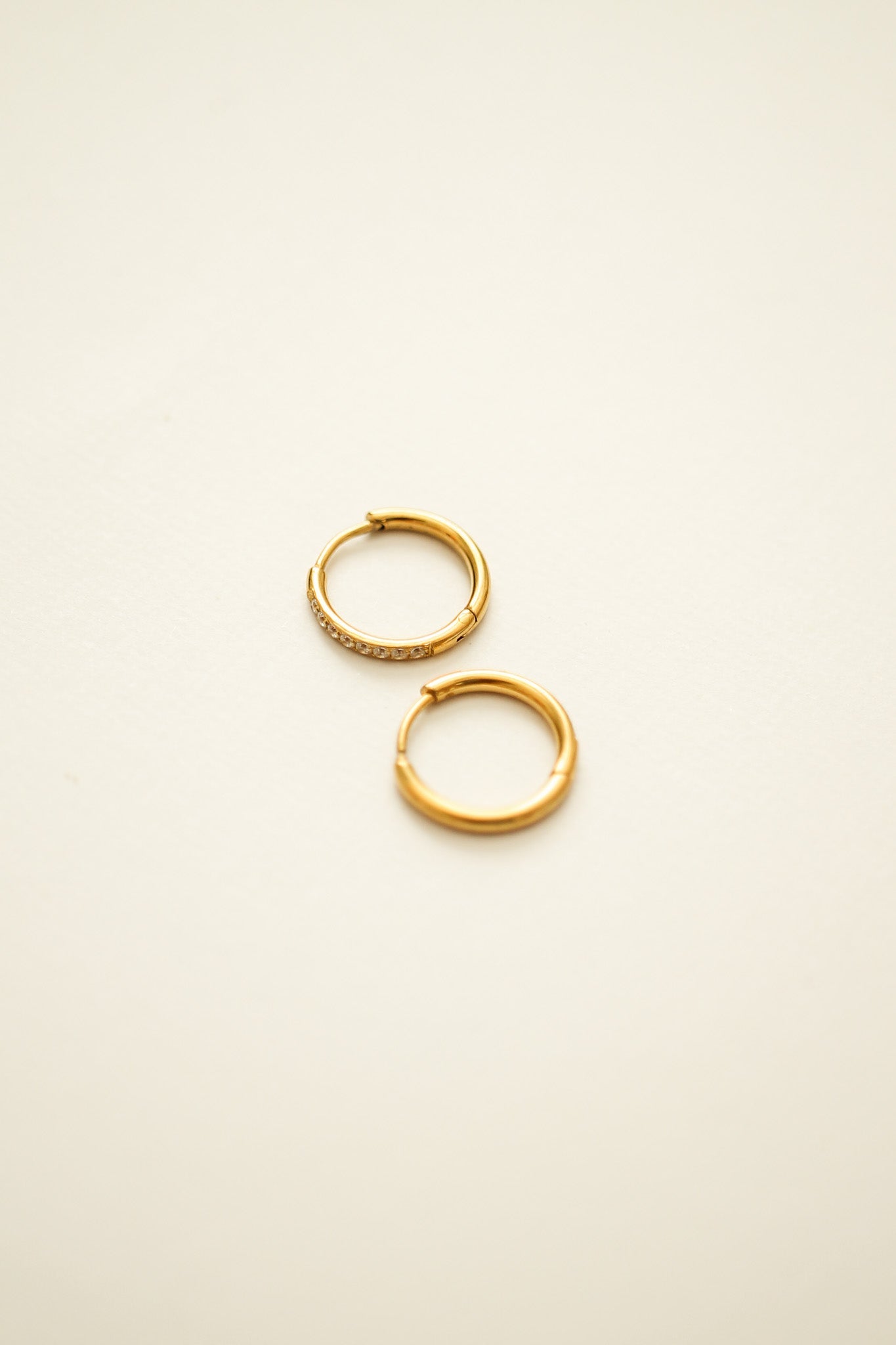 Two gold hoop earrings, one adorned with small sparkling stones, rest on a smooth, light-colored background, highlighting their elegant design and beauty.