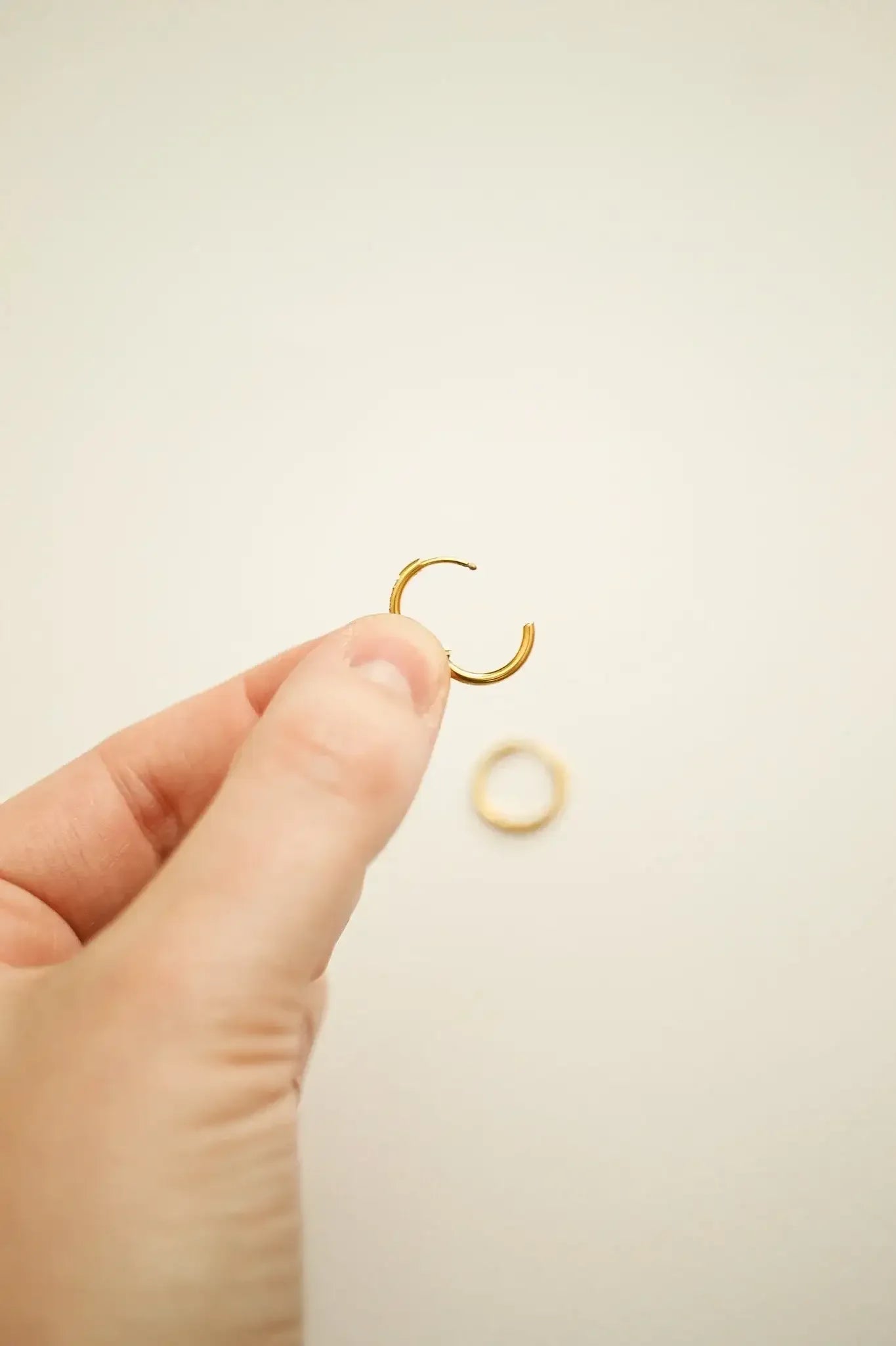 A hand holds a small gold hoop earring, while a second similar earring rests on a light-colored surface nearby, creating a simple and elegant jewelry display.