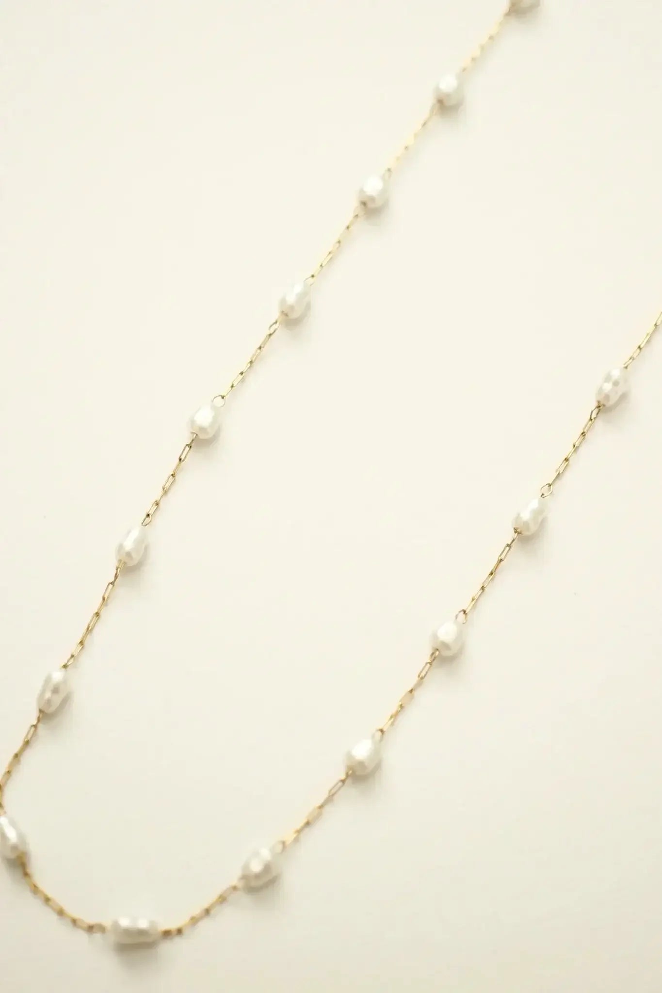 A delicate gold chain features irregularly shaped white pearls spaced along its length, elegantly lying flat against a soft, neutral background.