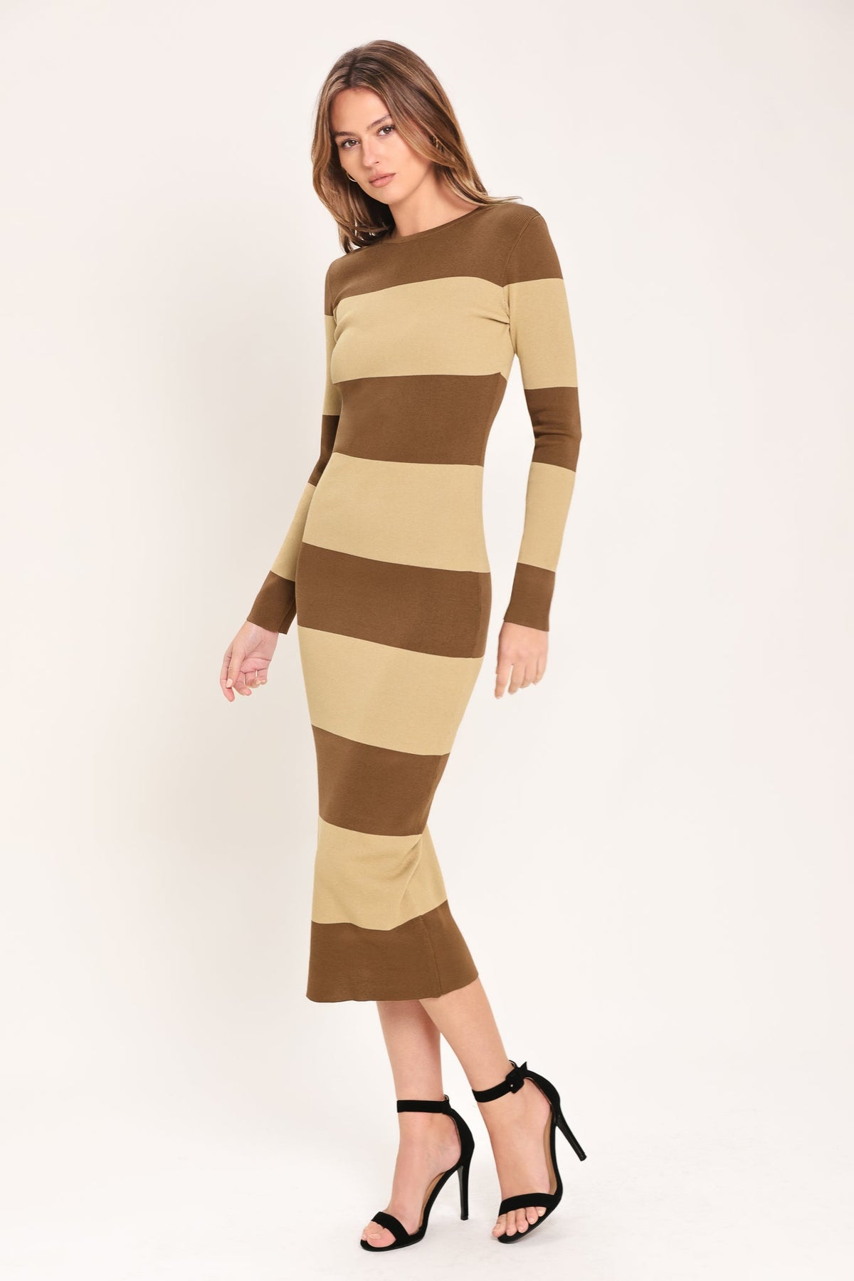 A woman modeling a long-sleeve bodycon midi dress with horizontal brown and beige stripes, paired with black high-heeled sandals, against a plain light background.