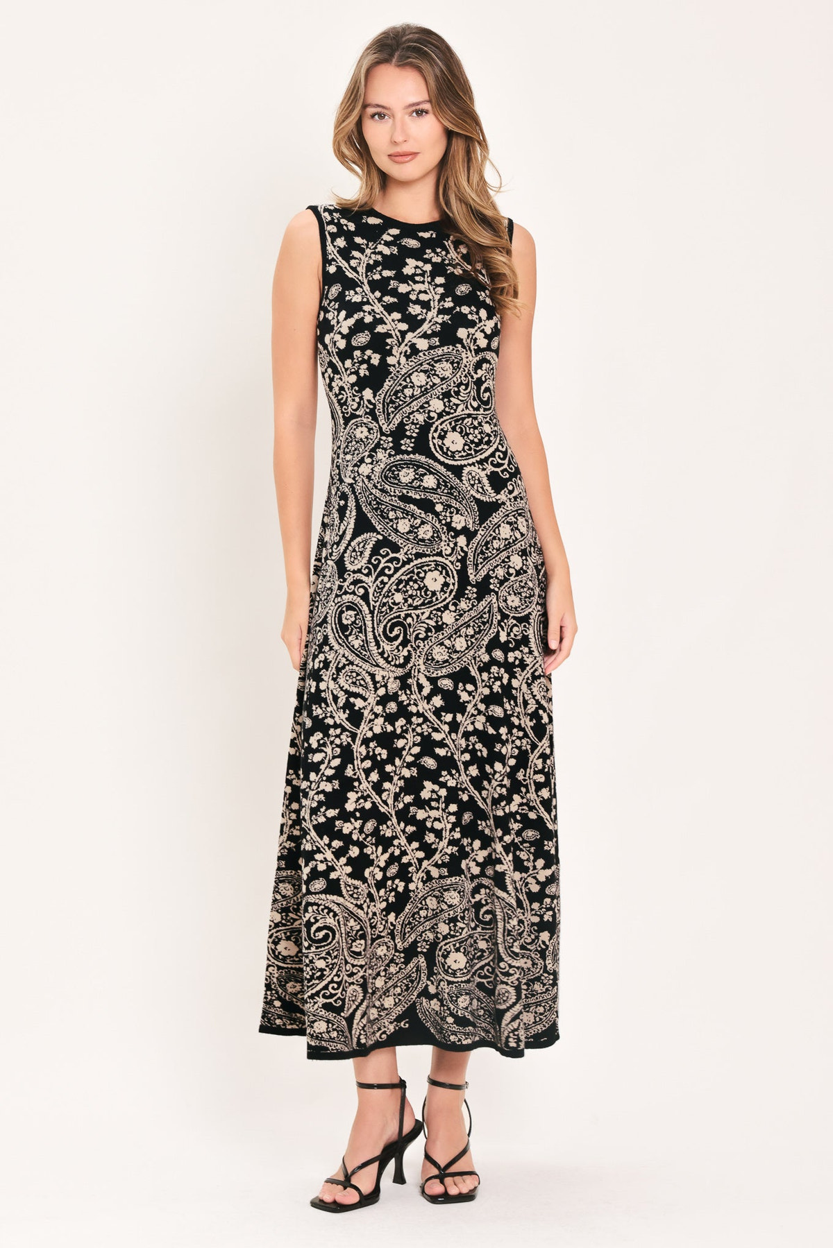 HAZEL PAISLEY EMBRACE JACQUARD SWEATER MIDI DRESS | Lady Black Tie | A sleeveless black maxi dress with an intricate beige paisley and floral pattern, worn by a model in black strappy heels against a plain off-white studio background.