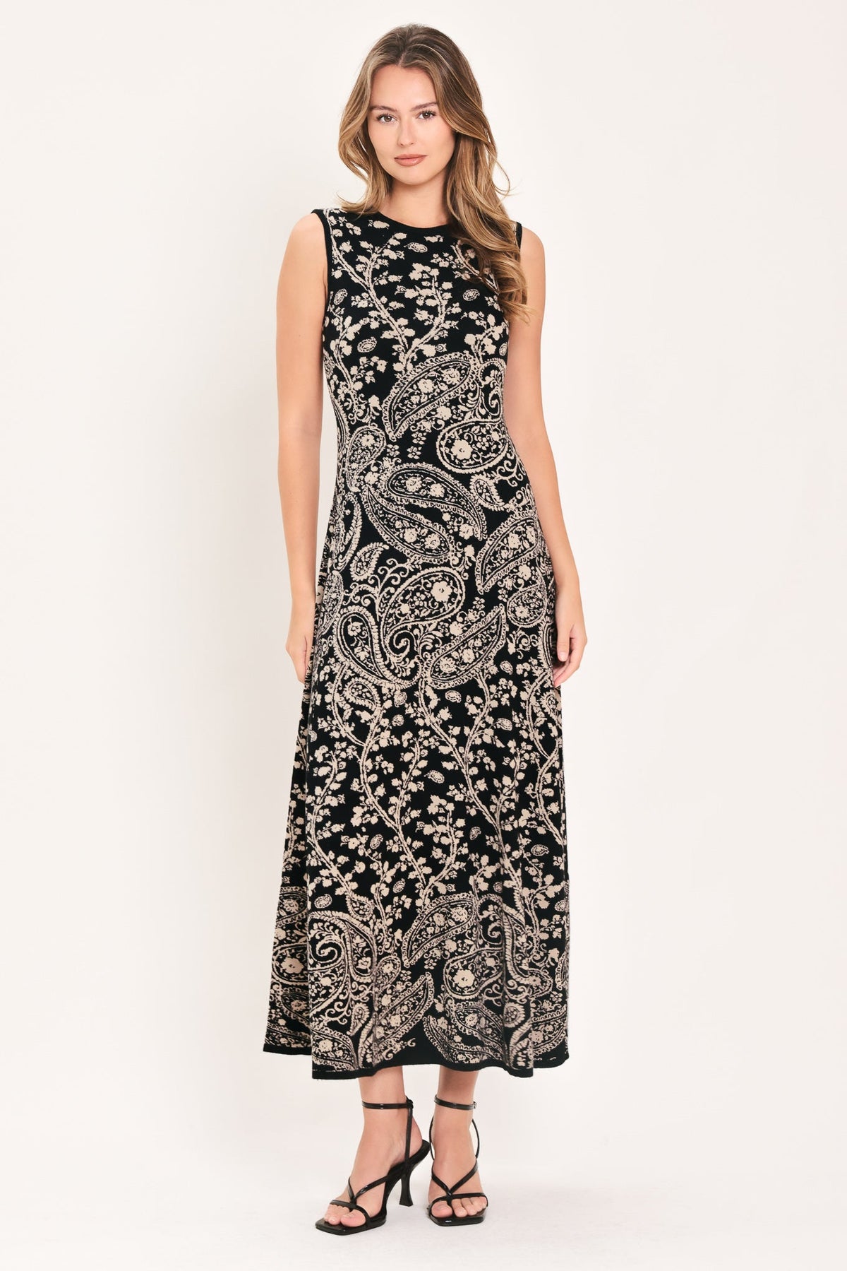 HAZEL PAISLEY EMBRACE JACQUARD SWEATER MIDI DRESS | Lady Black Tie | A sleeveless black maxi dress with an intricate beige paisley and floral pattern, worn by a model in black strappy heels against a plain off-white studio background.