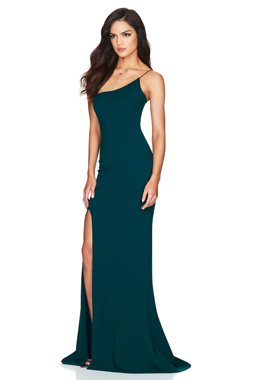 A woman stands confidently in a fitted, dark teal evening gown with a single shoulder strap and a thigh-high slit, showcasing elegance in a minimalist setting.