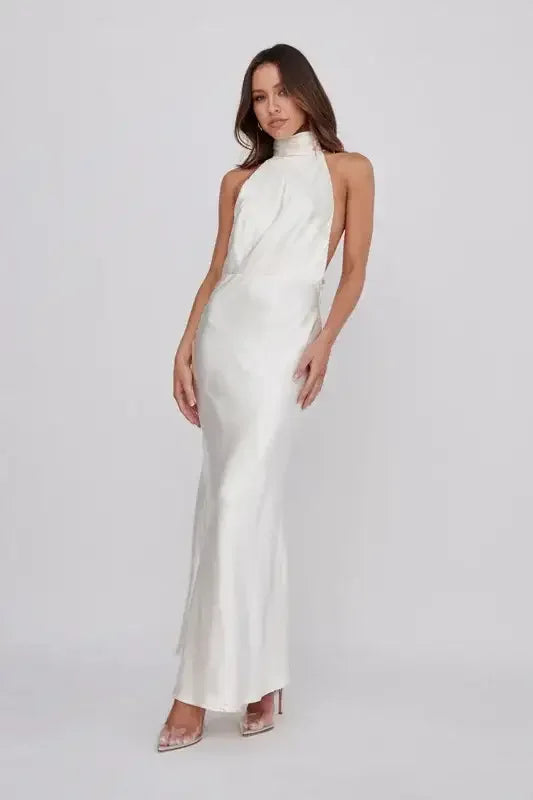 A model poses elegantly in a flowing, sleeveless, ivory dress with a high neck, showcasing a sleek silhouette against a plain, light gray background.