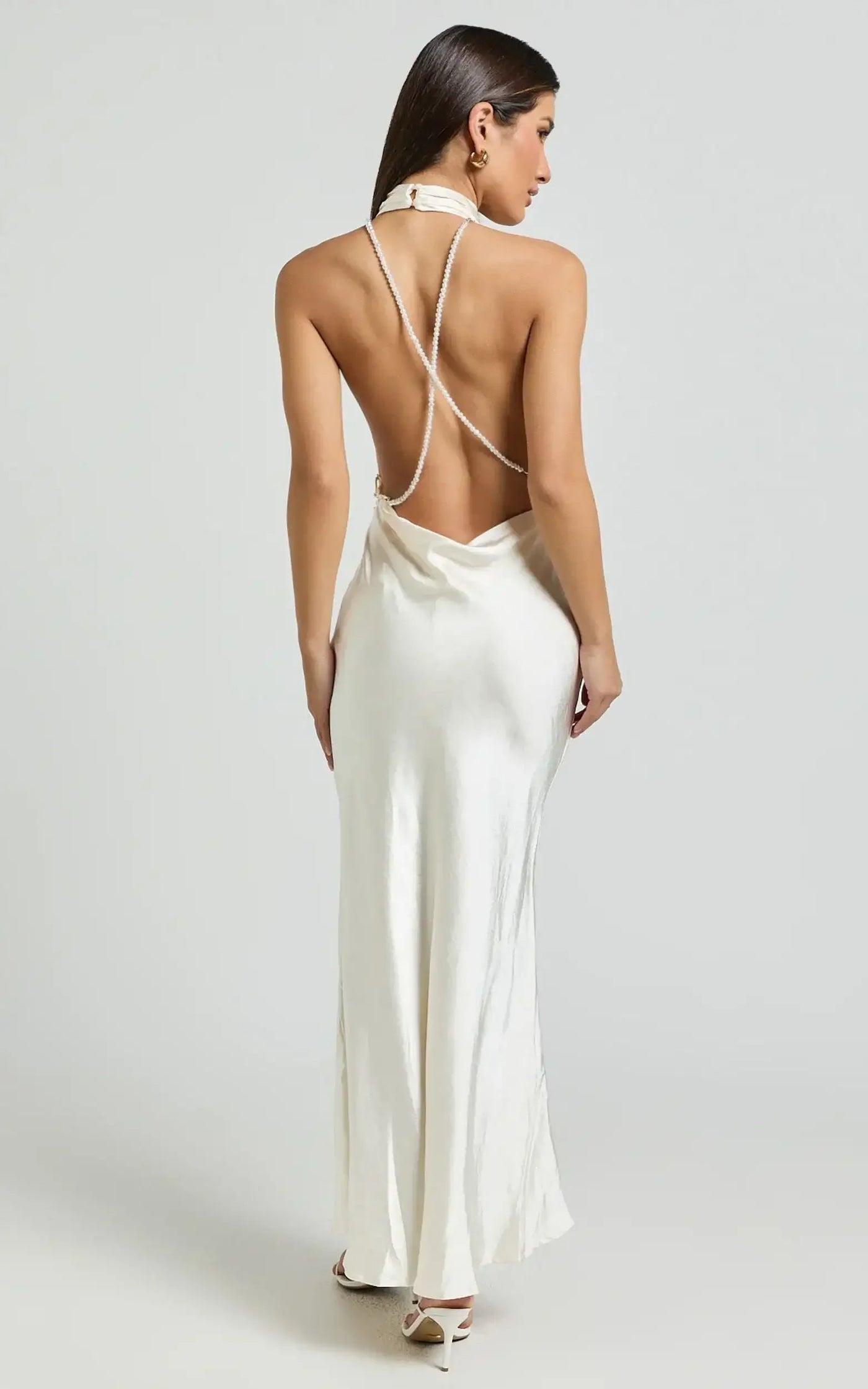 A woman wears a backless, ivory silk dress with intricate crisscross detailing, standing confidently against a light gray background. Her posture highlights the elegant design of the garment.