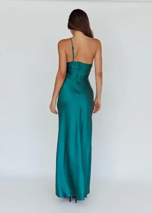 A woman stands with her back facing the viewer, wearing a long, teal satin dress with thin straps. The background is plain and neutral, emphasizing the elegant silhouette of the dress.