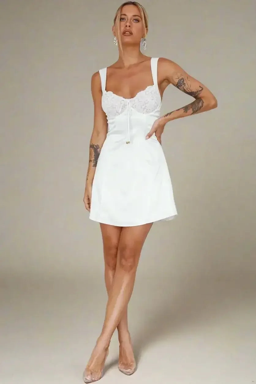 A woman stands confidently wearing a white dress with lace detailing and spaghetti straps, displaying tattoos on her arms. She poses in front of a light-colored backdrop and a textured floor.