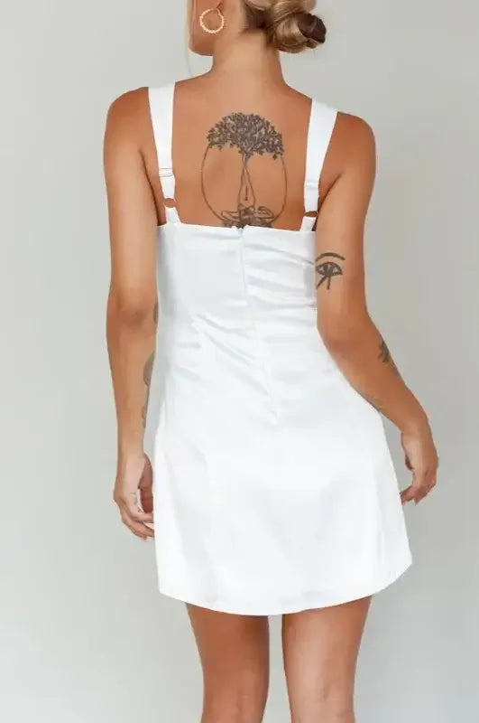 A person wearing a white dress stands against a plain background. The dress has thin straps and a fitted silhouette. A tree tattoo is visible on their back.