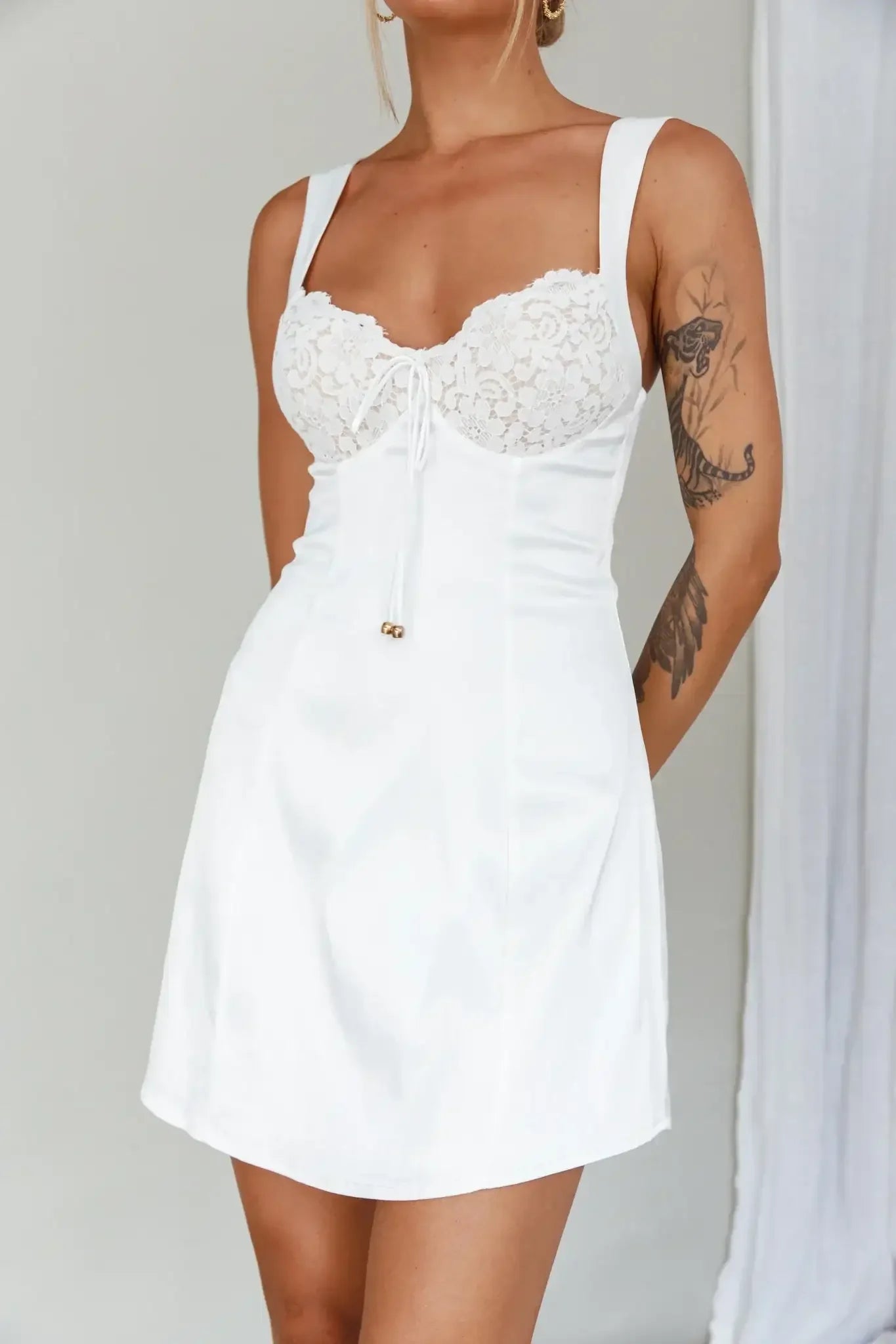 A woman wears a white dress with a lace top and straps, standing in front of a light-colored background. Her arm reveals a visible tattoo.