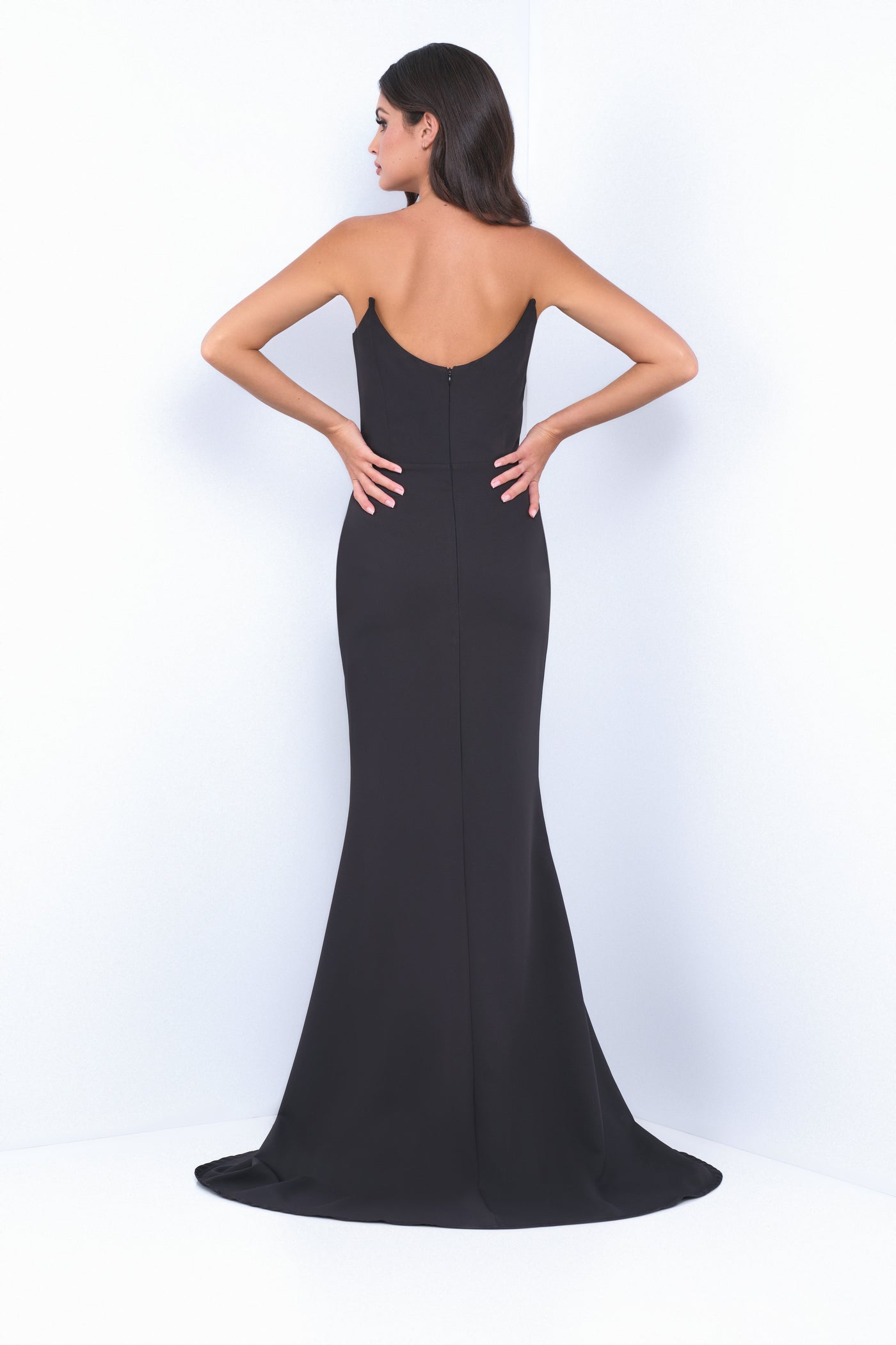A woman in a sleek black evening gown stands with her back facing the viewer, hands on her hips, in a minimalistic, bright setting with white walls.