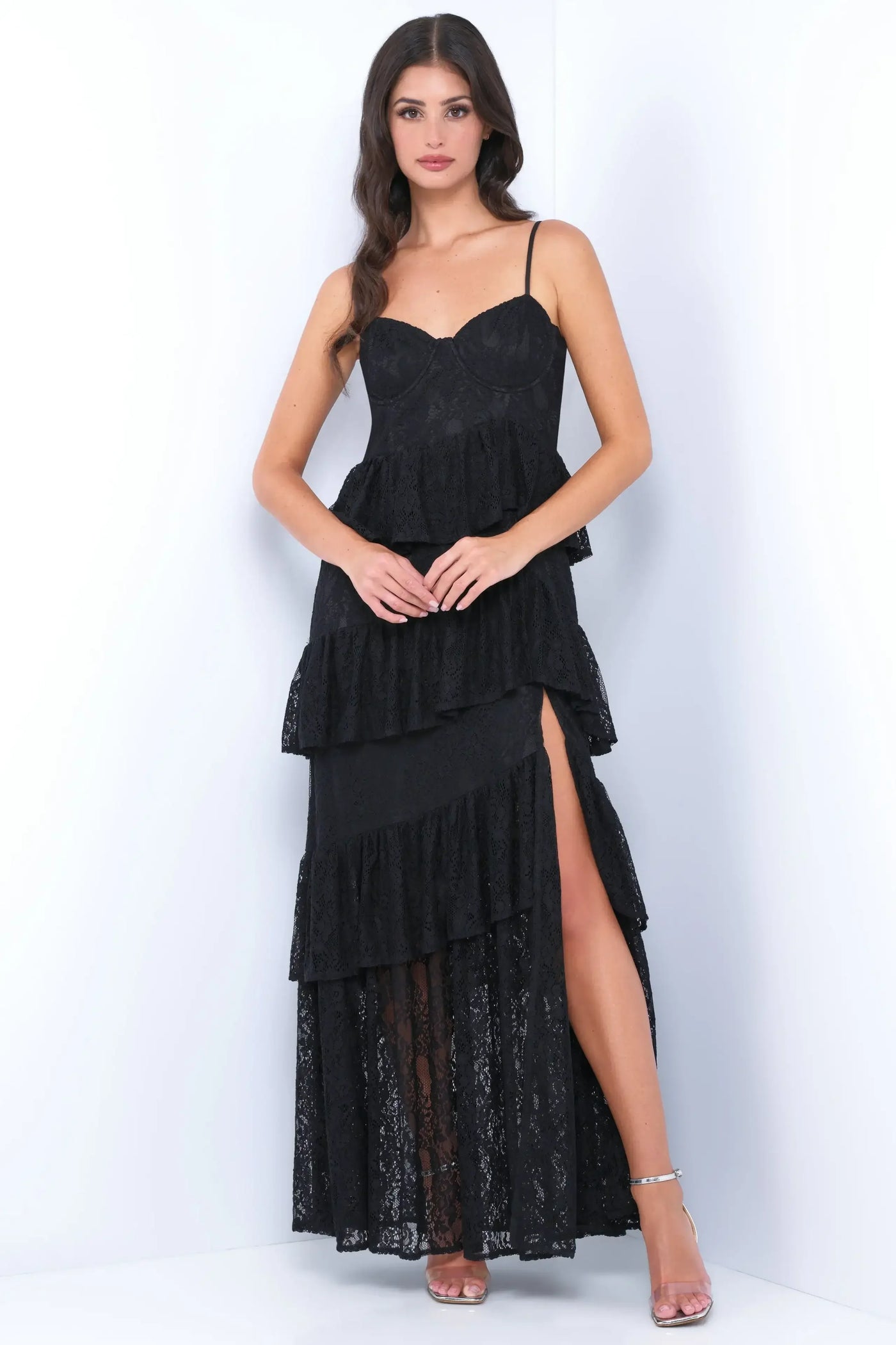 A model stands confidently wearing a black layered lace dress with a thigh-high slit. She has long hair, and the backdrop is a clean, white setting, enhancing the outfit's elegance.