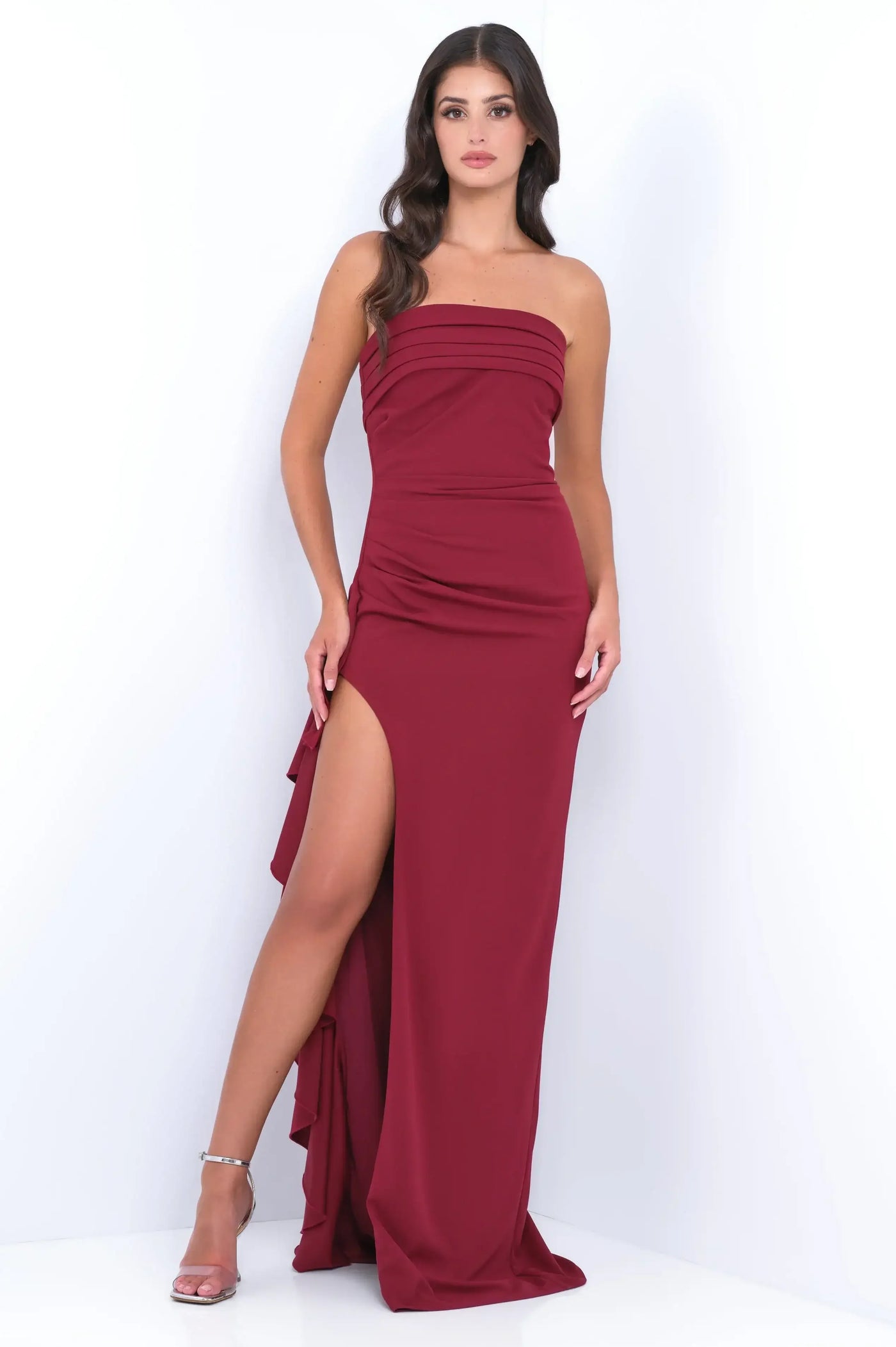 A woman stands confidently in a fitted, burgundy evening gown with a thigh-high slit. She poses against a bright, white background, showcasing the sleek design of her attire.