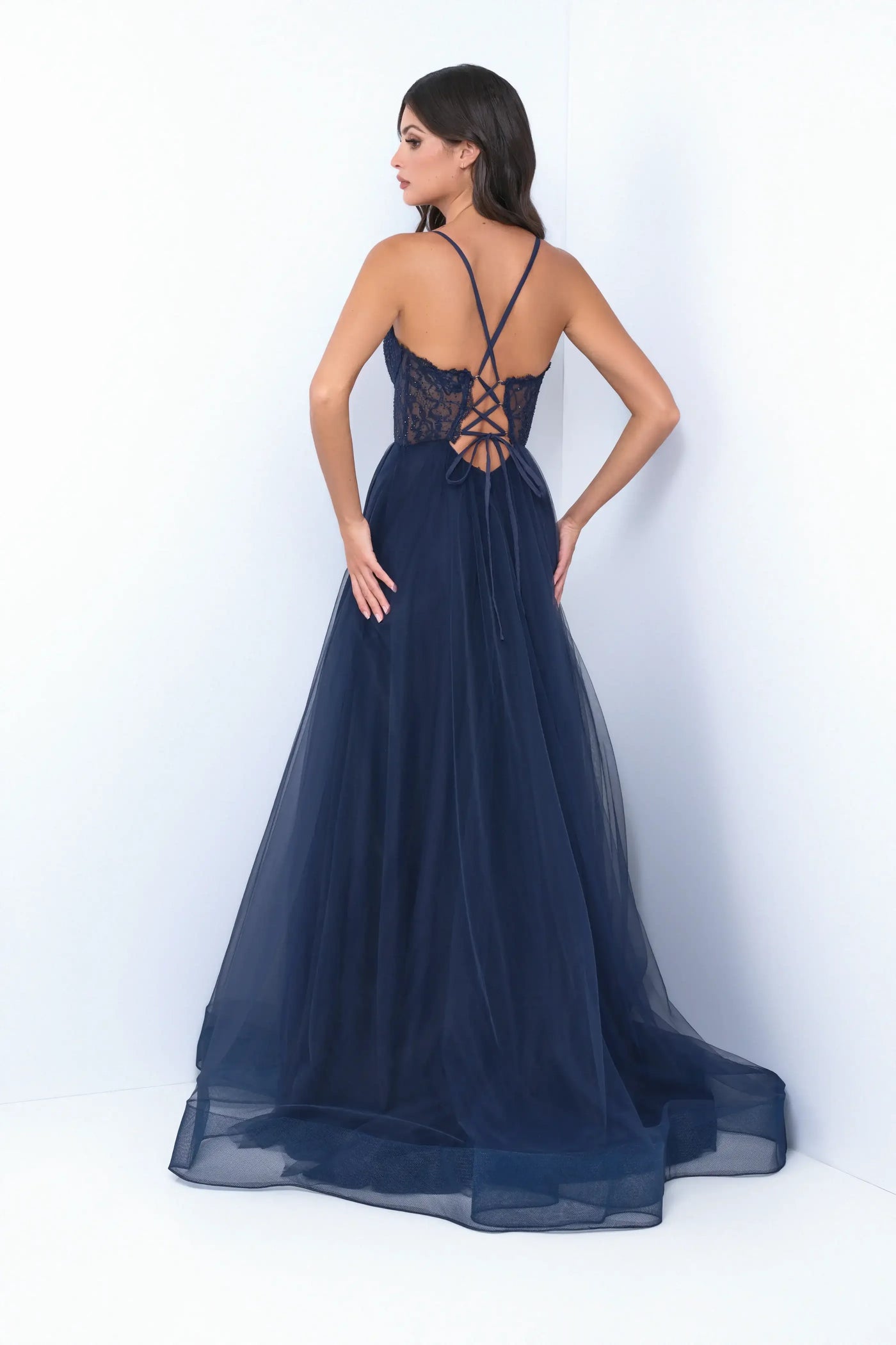 A woman stands gracefully, wearing a navy blue gown with intricate lace detailing and a flowing tulle skirt. She faces away, revealing a crisscross back design in a minimalist setting.