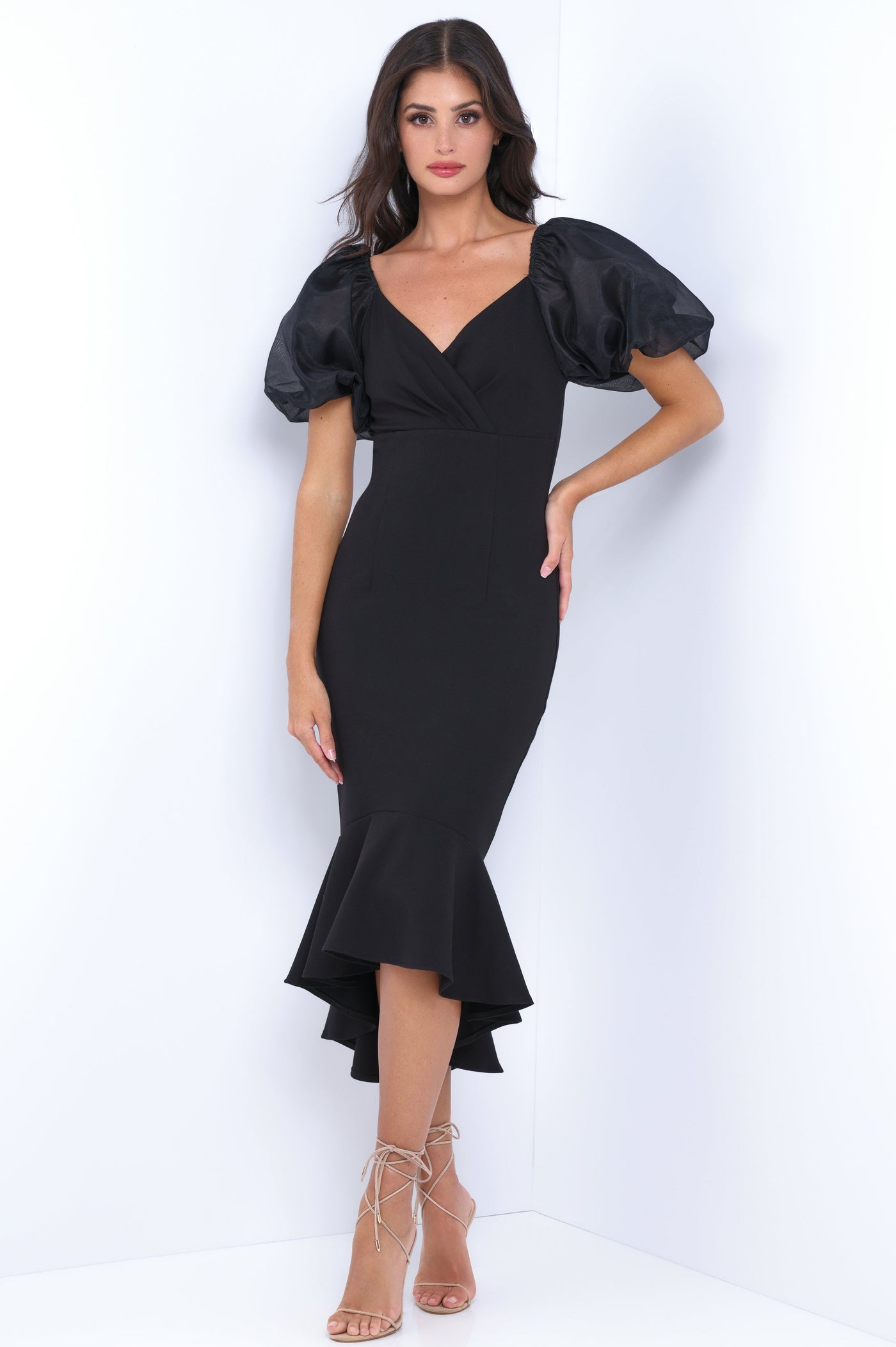 A woman stands in a fitted black dress with puffed sleeves and a flared hem. She poses gracefully against a white wall, showcasing her elegant outfit and strappy heels.