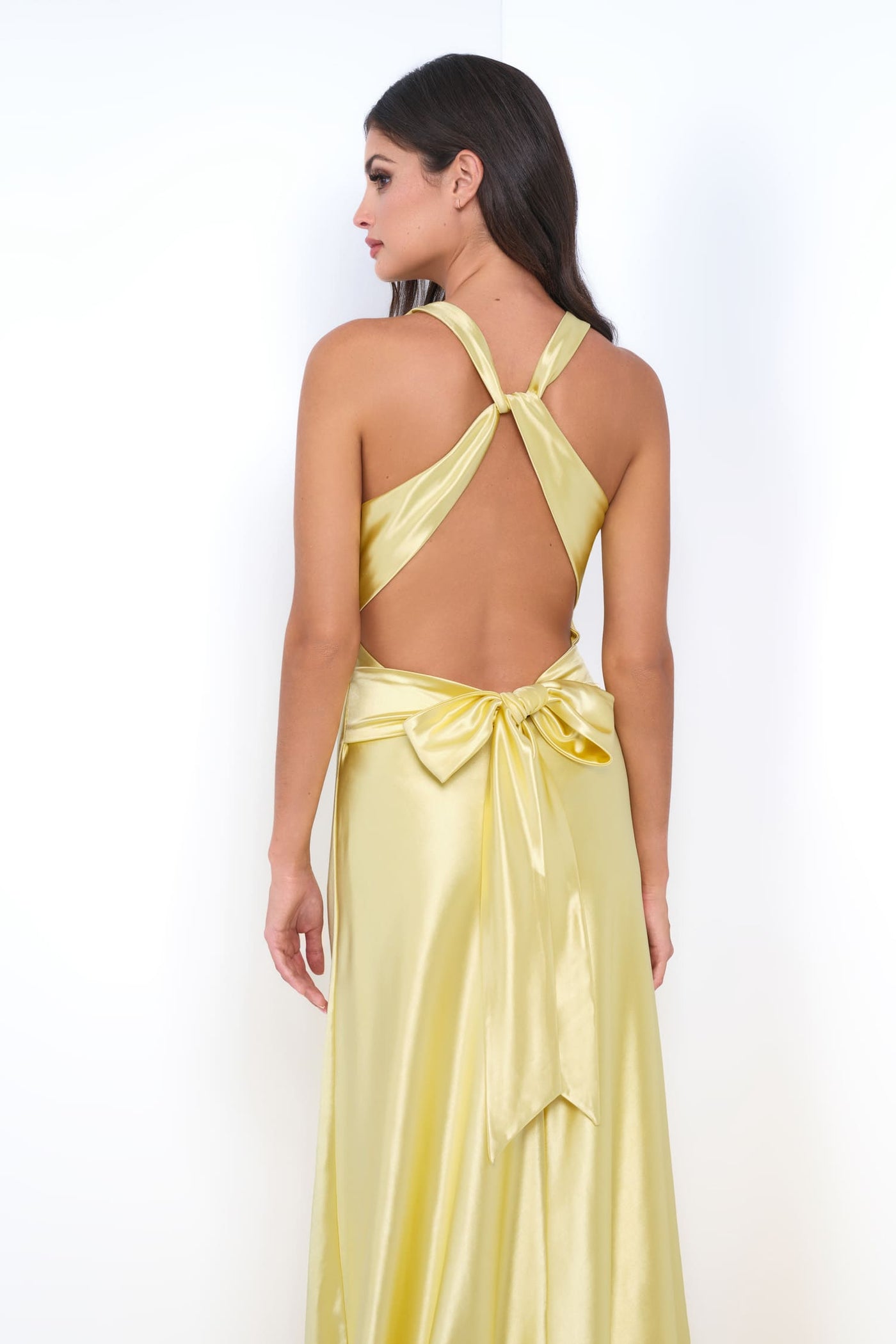 A woman modeling a yellow satin maxi dress from the back, featuring a halter-style open back with crossed straps and a large bow at the waist, set against a plain white background.