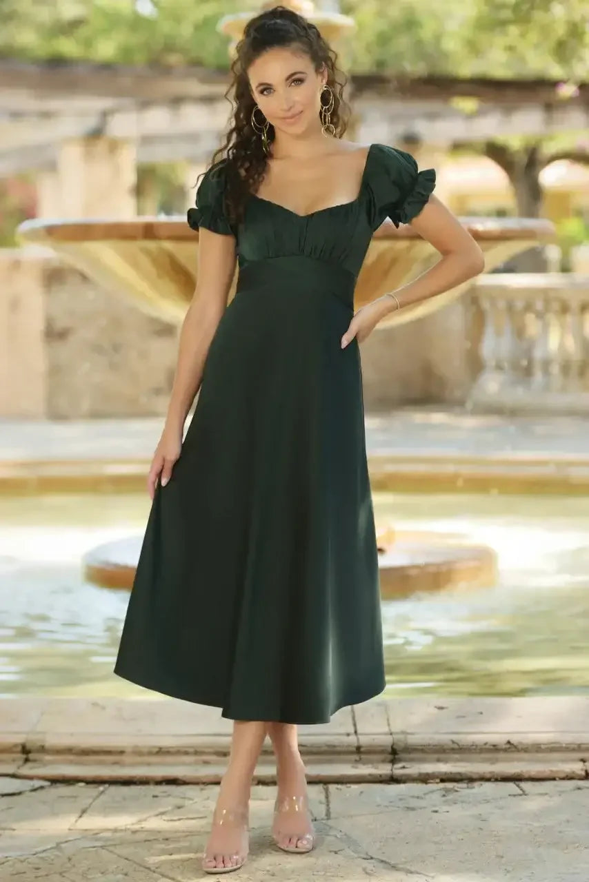 A woman stands by a fountain, wearing a dark green, mid-length dress with puff sleeves. She poses confidently, surrounded by a sunny outdoor garden with soft greenery.