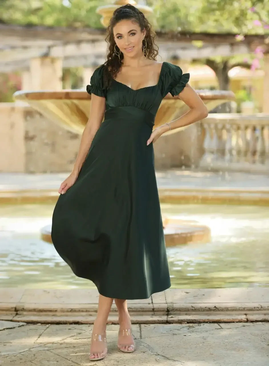 A woman in a green dress stands gracefully by a fountain, smiling. She wears hoop earrings and strappy sandals, surrounded by a serene outdoor setting with greenery and stone architecture.