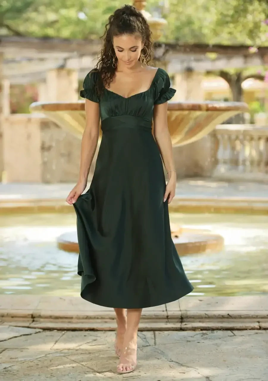 A woman in a dark green dress gently lifts the hem while gazing down. She stands near a fountain in a sunlit, lush garden setting, with soft greenery in the background.