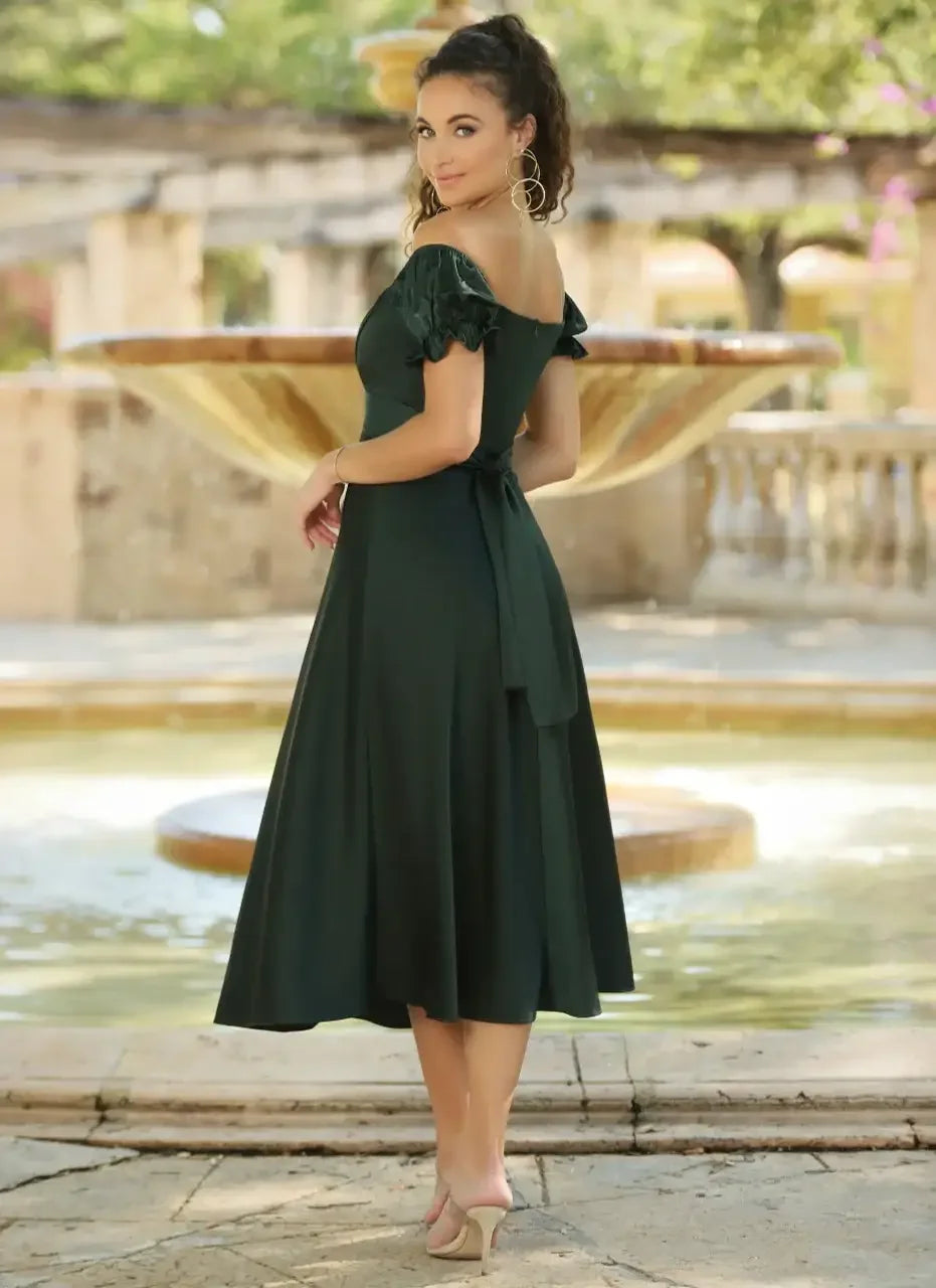 A woman wearing an off-shoulder green dress stands gracefully by a fountain, her body angled slightly to the side, in a lush garden setting with soft sunlight filtering through the trees.