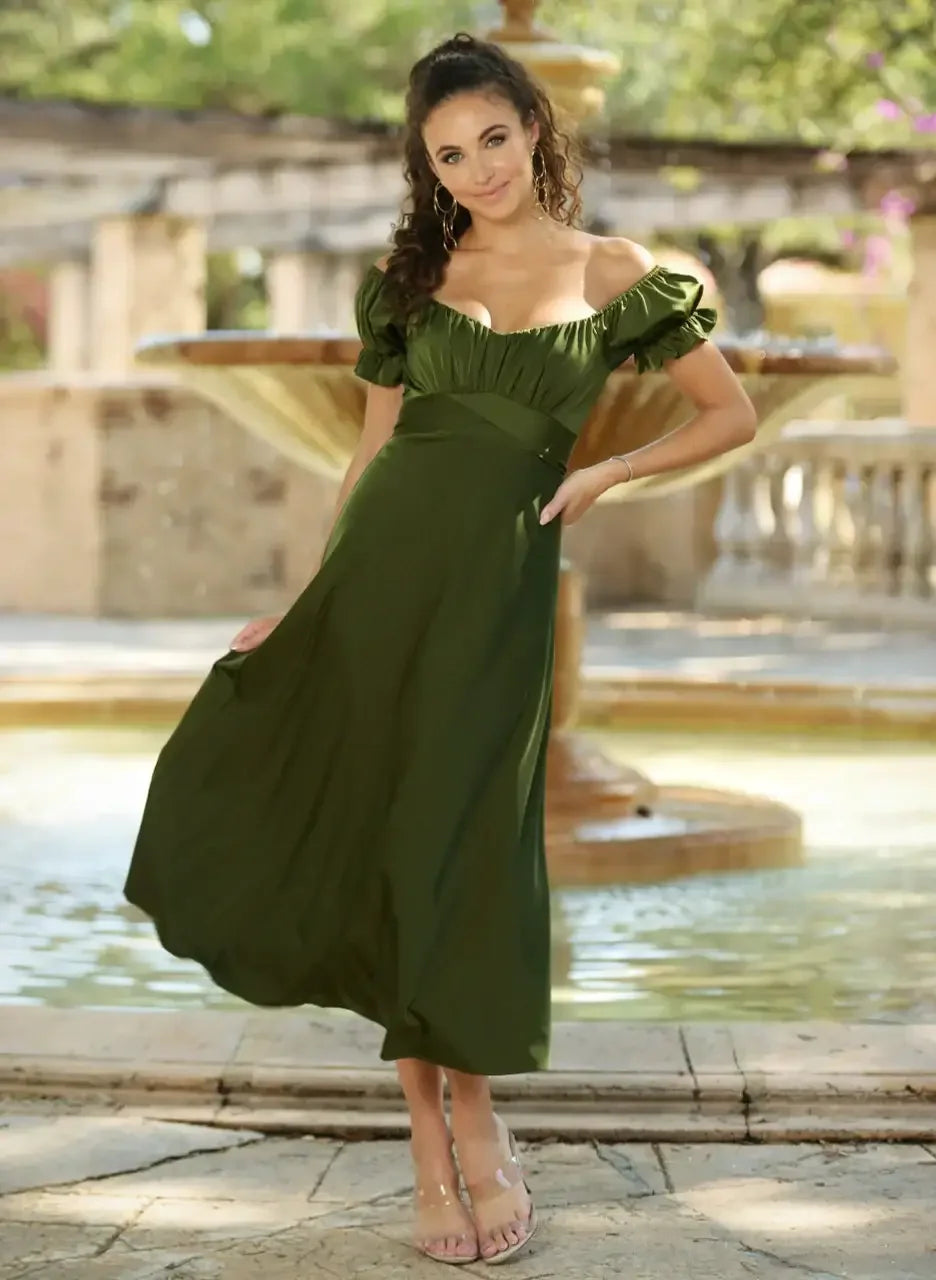 A woman stands gracefully in a flowing dark green dress near a fountain, smiling. The setting is an idyllic outdoor space, surrounded by soft sunlight and greenery.