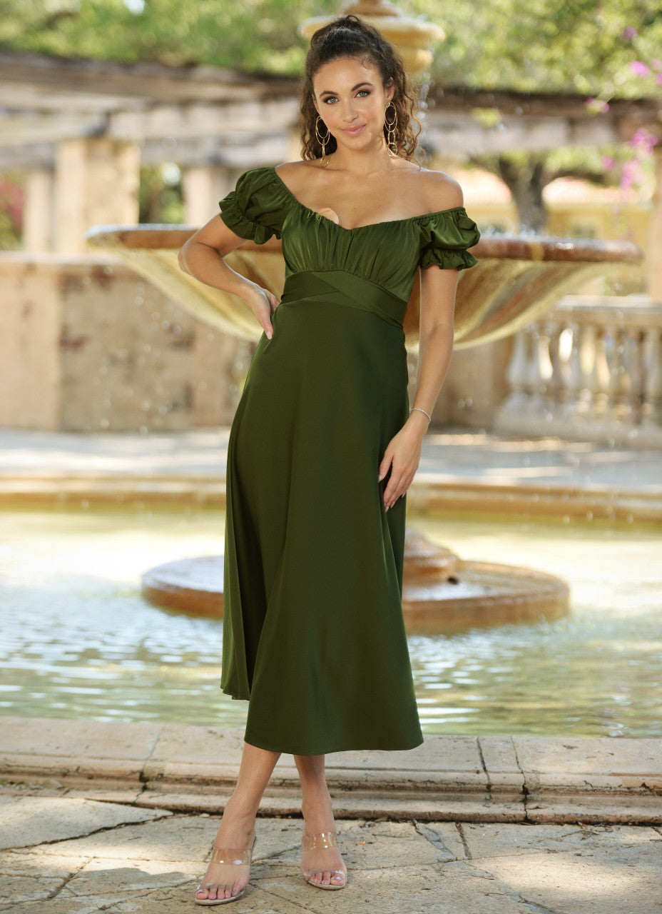A woman in a green off-the-shoulder dress stands confidently by a fountain, posing with a smile. The background features lush greenery and a stone setting, suggesting an outdoor venue.