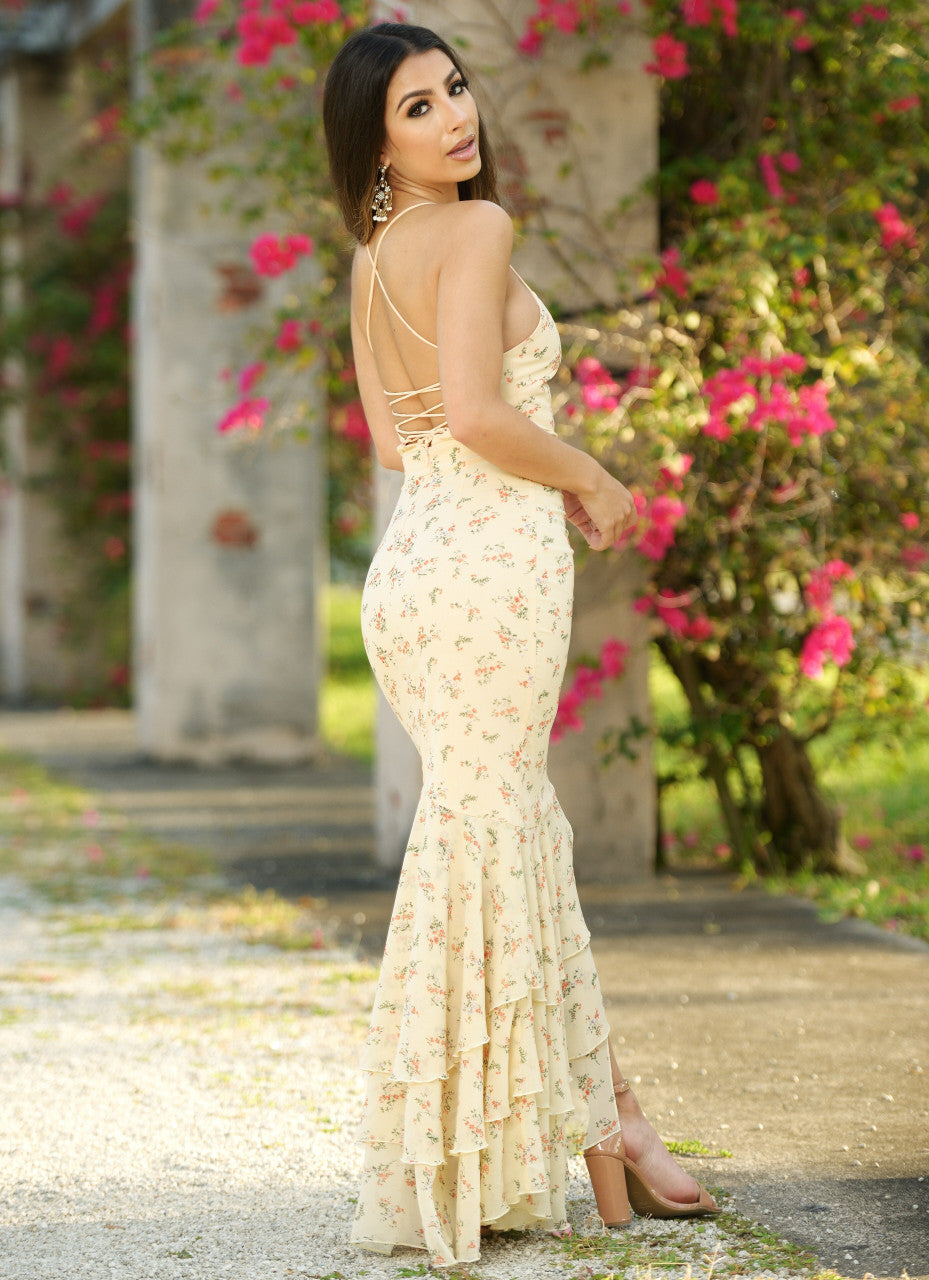 A young woman wearing a fitted, floral-patterned gown with a ruffled hem stands gracefully in a garden, showcasing the dress's trailing back straps against a backdrop of vibrant pink flowers.