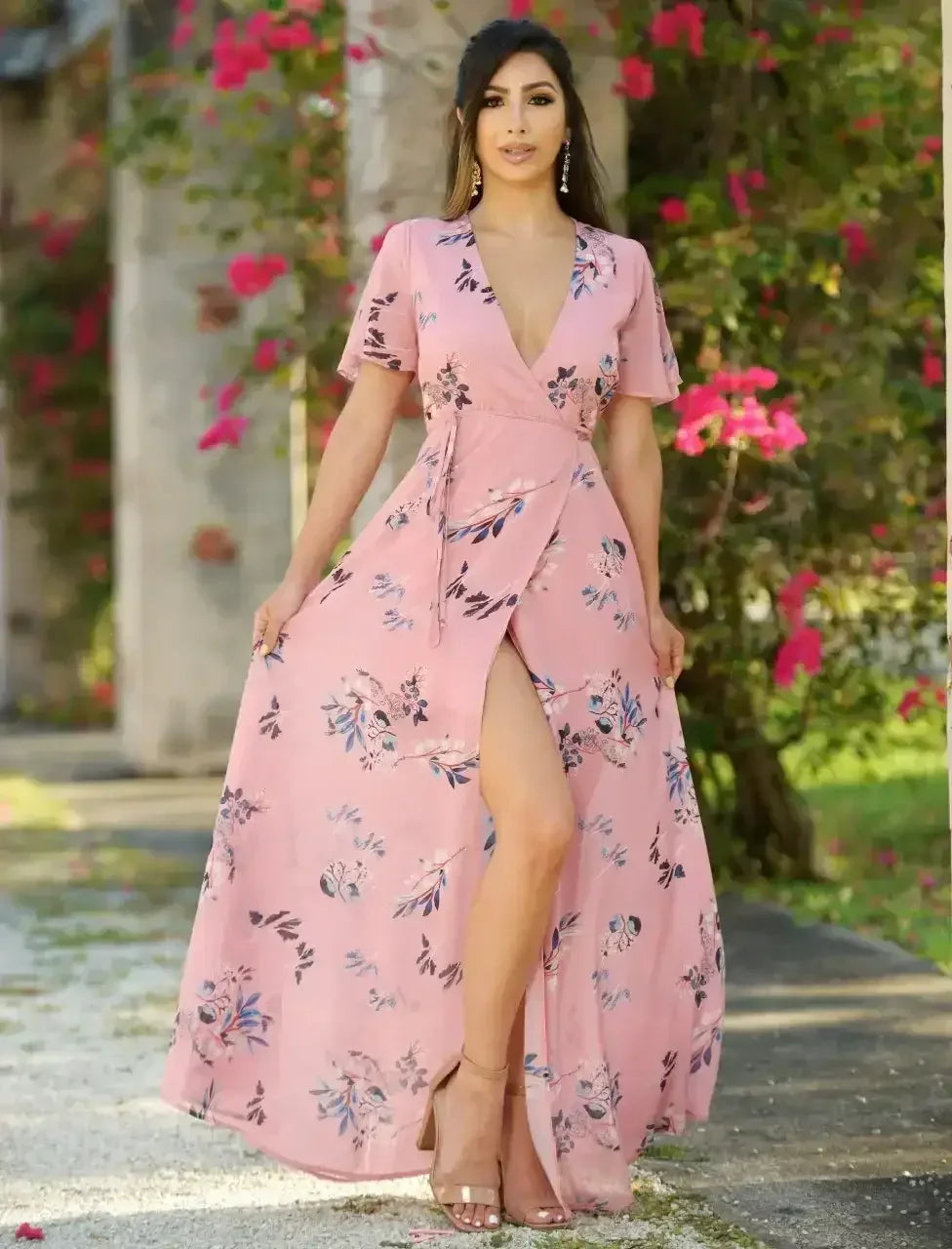 A woman in a floral pink maxi dress with a thigh-high slit is posing confidently. She stands amidst vibrant greenery and blooming flowers, showcasing a natural, sunny outdoor setting.