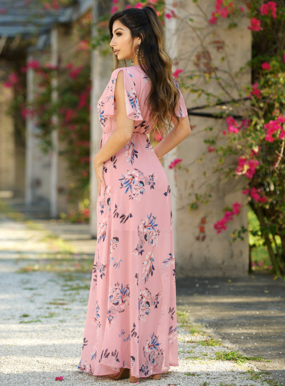 A woman poses confidently in a floral pink maxi dress with flutter sleeves. She stands amidst a garden, surrounded by blooming flowers against a backdrop of stone columns and pathways.
