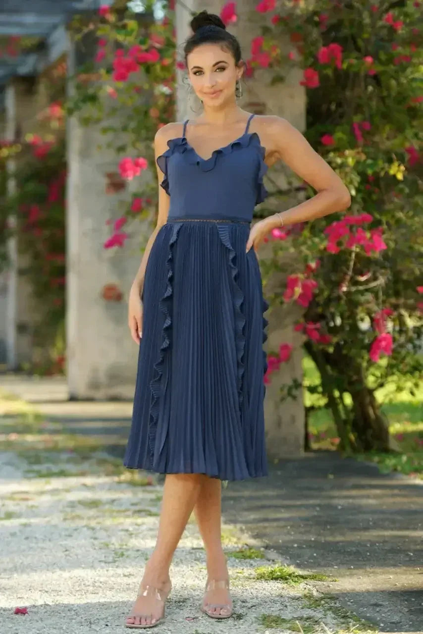 A woman stands gracefully in a navy dress with ruffled details and pleats. She poses against a backdrop of vibrant pink flowers and stone architecture in a sunny outdoor setting.