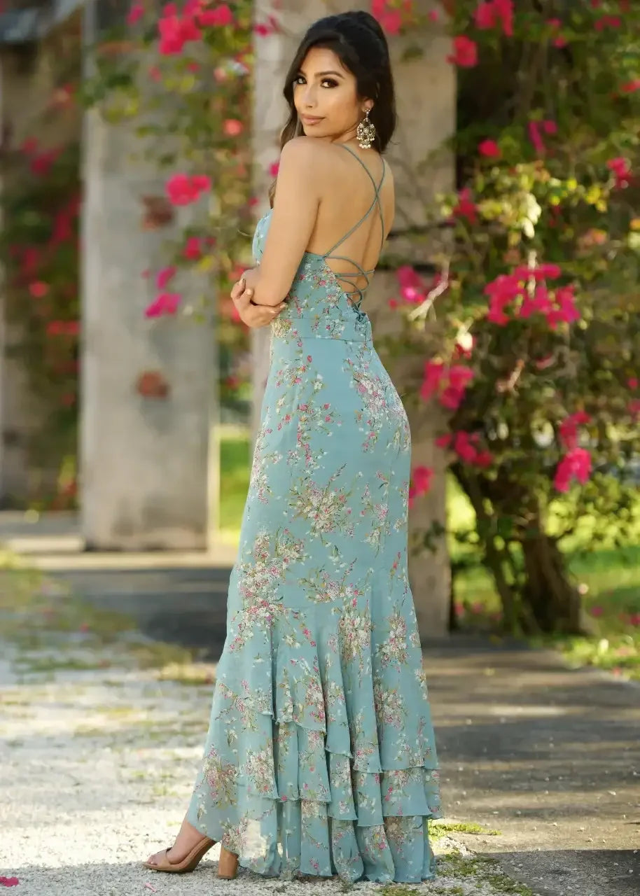 A woman in a floral, light blue dress stands gracefully, facing sideways. She poses amidst vibrant pink flowers and greenery, highlighting a sunny, outdoor setting.