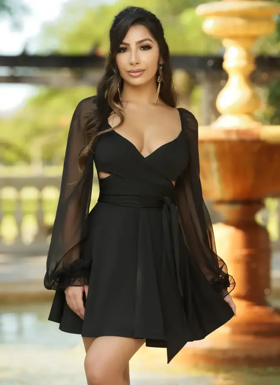 A woman stands confidently in a flowing black dress with sheer sleeves, posing by a decorative fountain in a lush garden, radiating elegance and beauty.