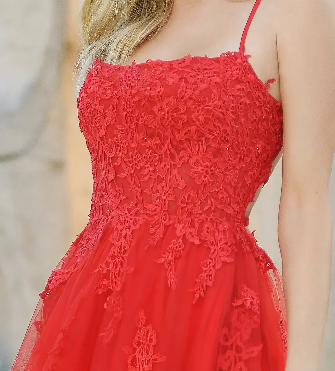 A red, lace-embroidered dress features delicate floral patterns and a flowing skirt. The wearer, with shoulder-length hair, stands in a softly blurred outdoor setting, exuding elegance.