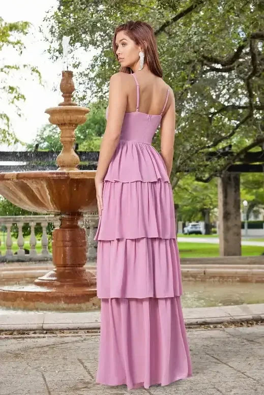 A woman wearing a layered, lavender gown stands near a fountain, facing away, with a garden backdrop featuring lush trees and a stone walkway surrounding the water feature.