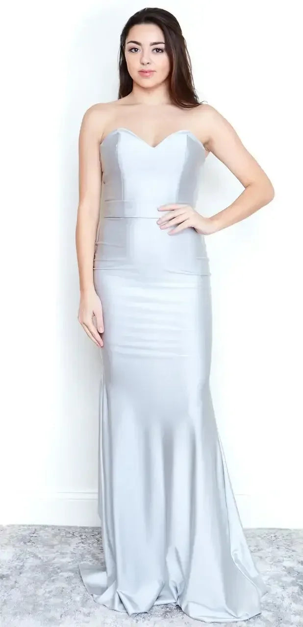 A person stands wearing a form-fitting, strapless silver gown that flows to the floor. They are positioned against a plain white background, showcasing the dress's elegant design.