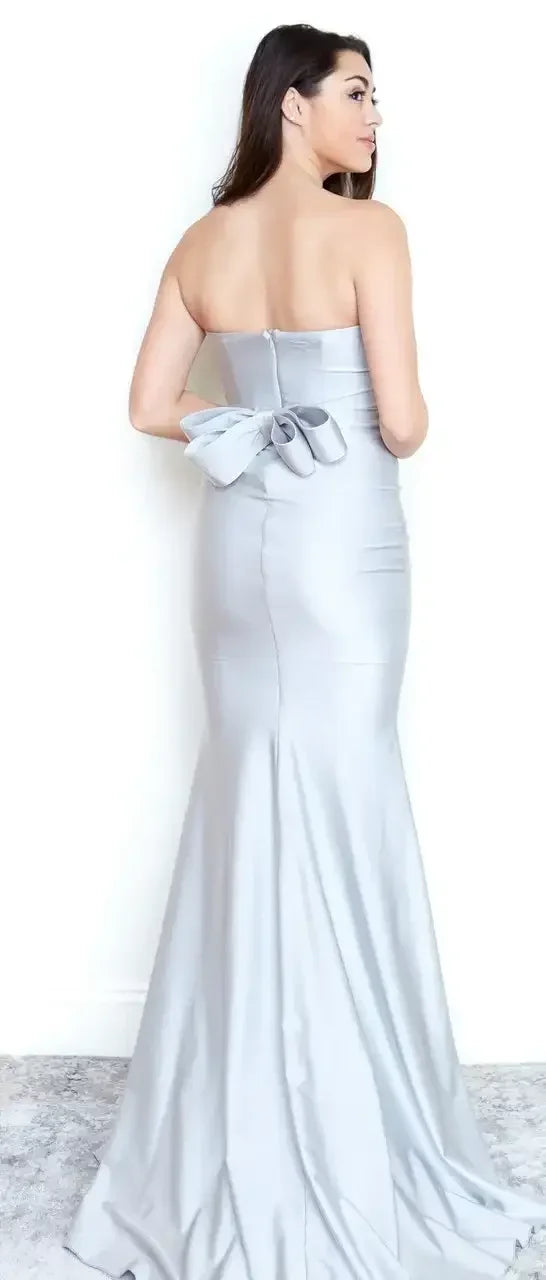 A woman is standing with her back to the viewer, wearing a fitted strapless silver gown that flares at the bottom. The dress features a large bow at the waist, set against a plain backdrop.