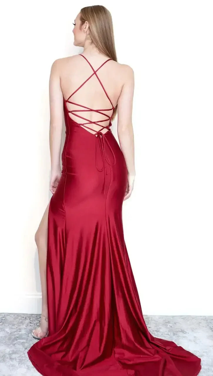 A woman wears a form-fitting, deep red satin evening dress with a thigh-high slit and crisscrossed back straps, standing against a plain white wall with a soft grey rug beneath her feet.