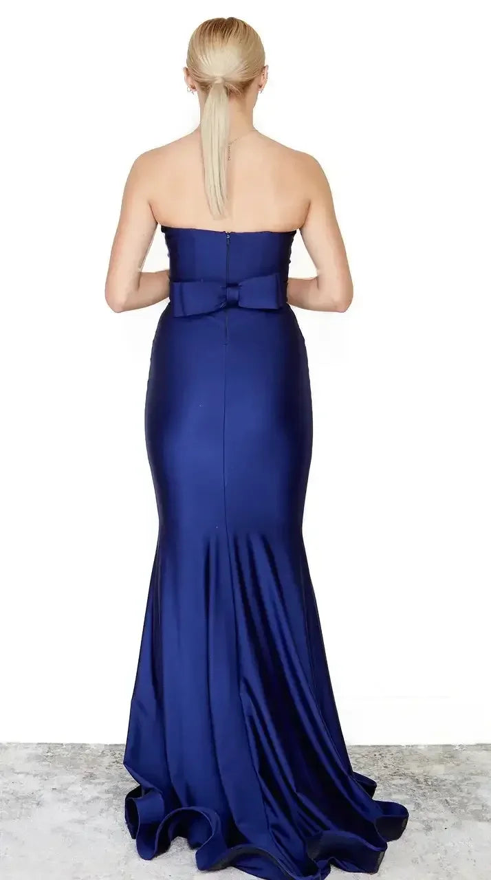 A woman stands facing away, wearing a strapless navy blue gown with a fitted bodice and a flared hem. The gown features a back zipper and a bow accent. A neutral background and textured floor are visible.