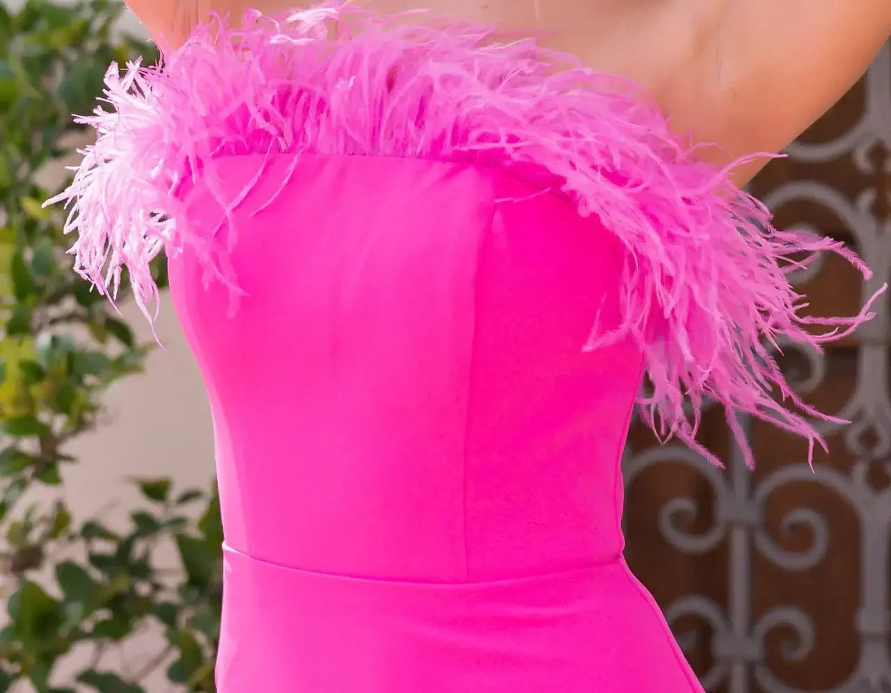 A vibrant pink dress with feather trim is highlighted, showcasing its bold color and playful texture. The background features soft greenery, creating an elegant outdoor setting.