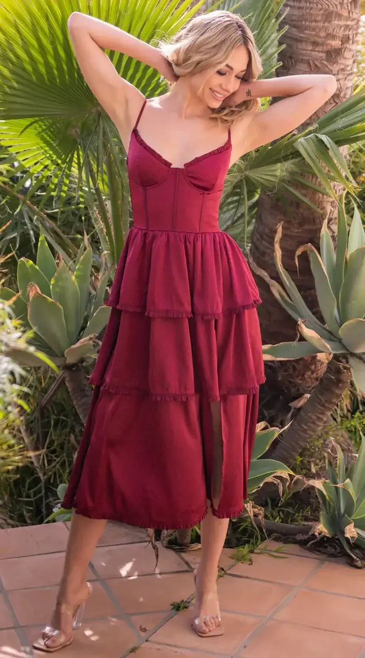 A woman in a flowing, tiered maroon dress is playfully adjusting her hair while standing on a tiled patio surrounded by lush green plants and palms. Her smile suggests joy.