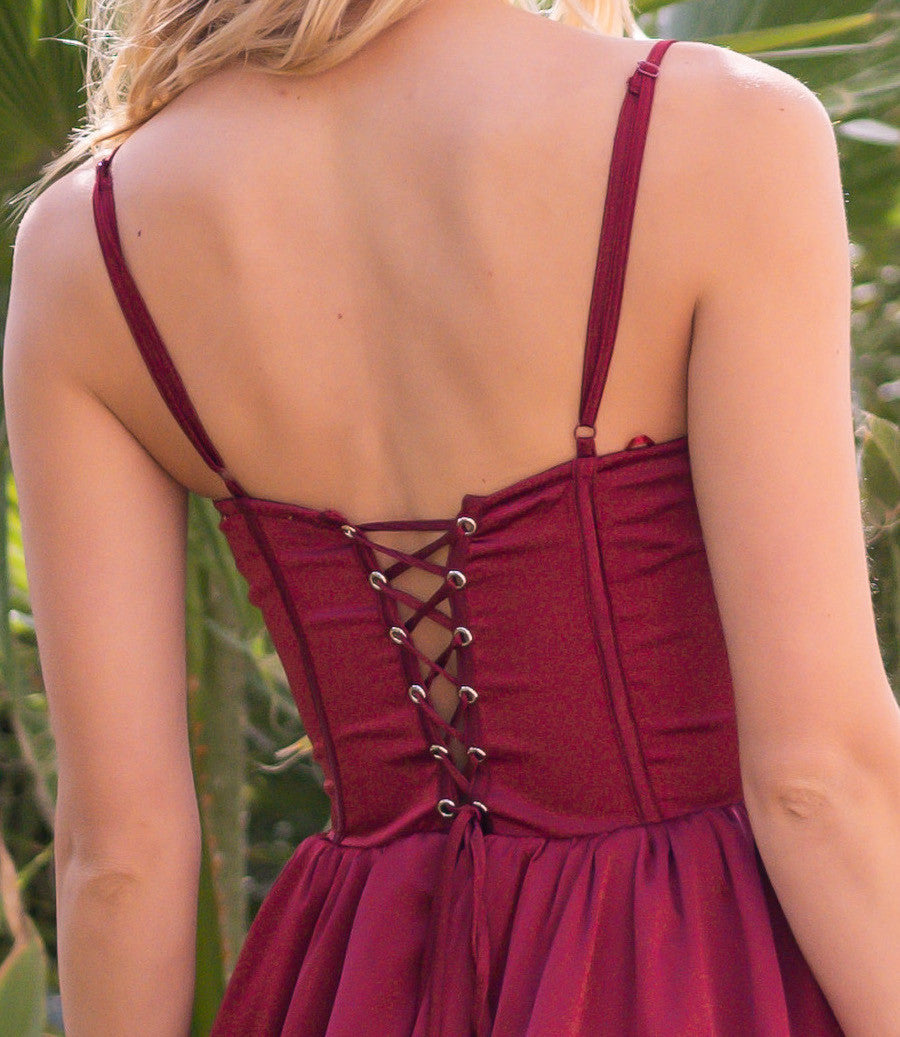 A burgundy dress features a lace-up back design with crisscrossed ribbons, highlighting a fitted bodice and flowing skirt. The background includes lush greenery, creating a vibrant, natural setting.