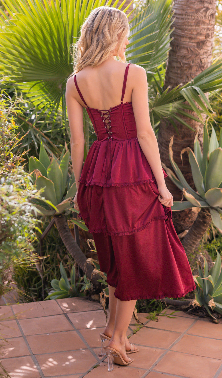 A woman in a burgundy dress with layered, ruffled fabric stands outdoors. She faces away, showcasing the lace-up back, surrounded by lush green plants and tiled flooring.