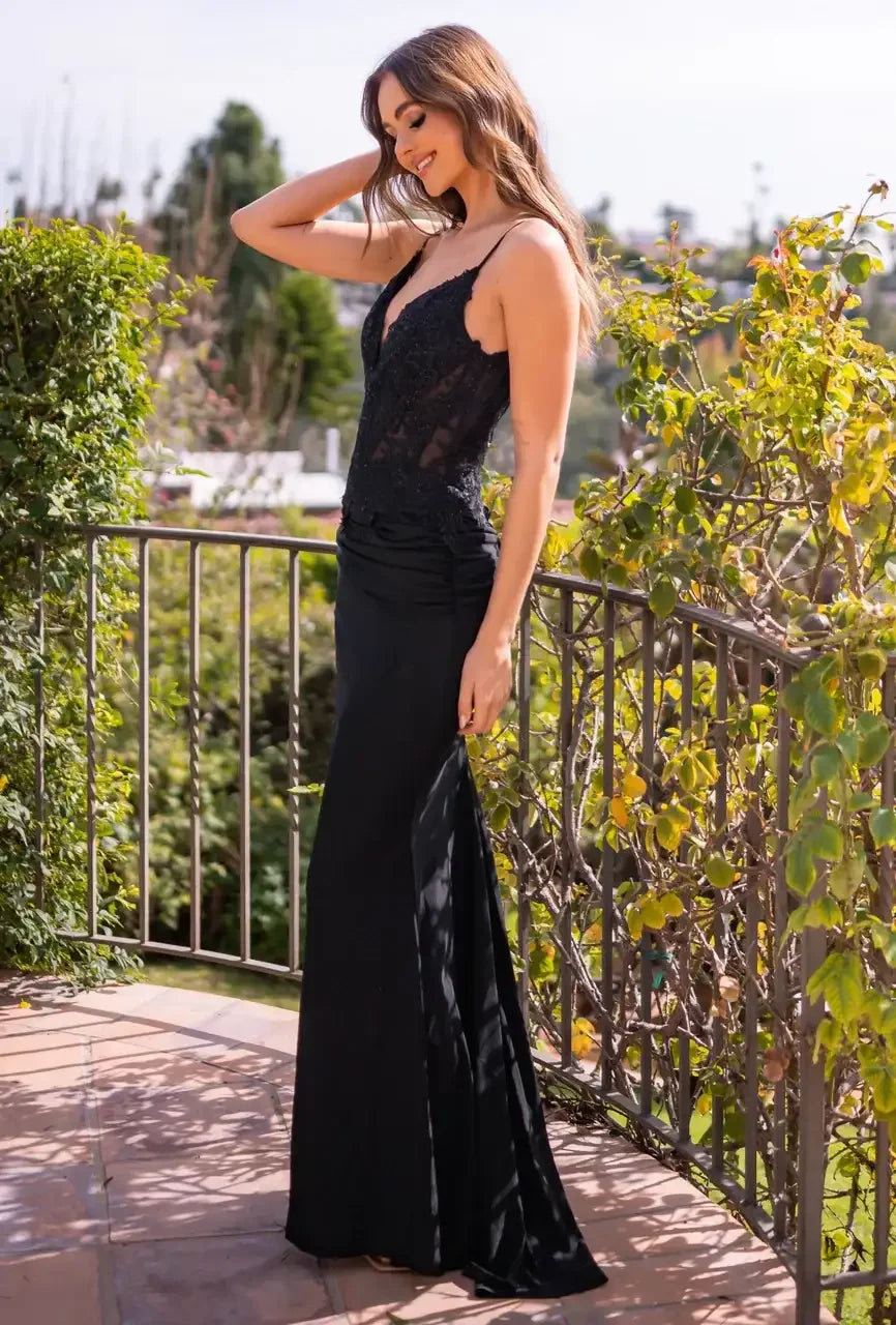 A woman in a black elegant dress stands gracefully on a balcony, smiling and touching her hair, surrounded by lush greenery under a clear sky.