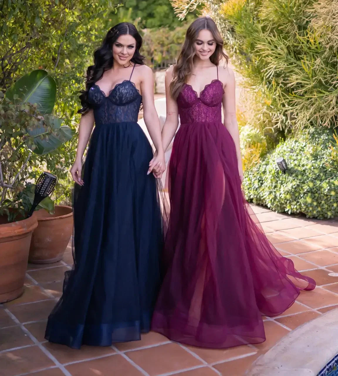 Two women are holding hands, wearing elegant, flowing dresses—one in navy blue and the other in burgundy. They are walking through a lush, greenery-filled outdoor area.