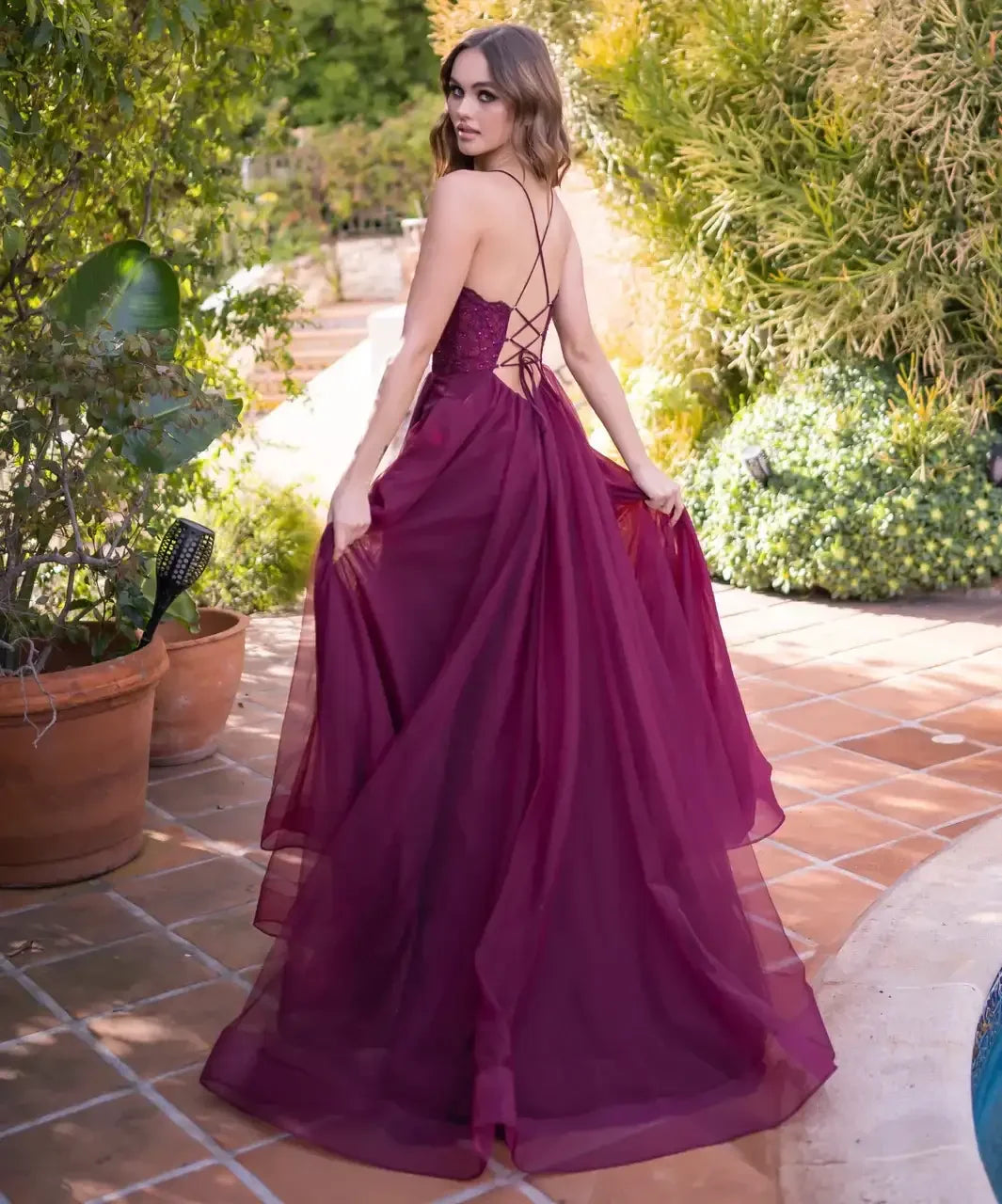 A young woman in a flowing burgundy gown stands gracefully, looking over her shoulder. She is surrounded by greenery and terracotta pots, set near a poolside area.